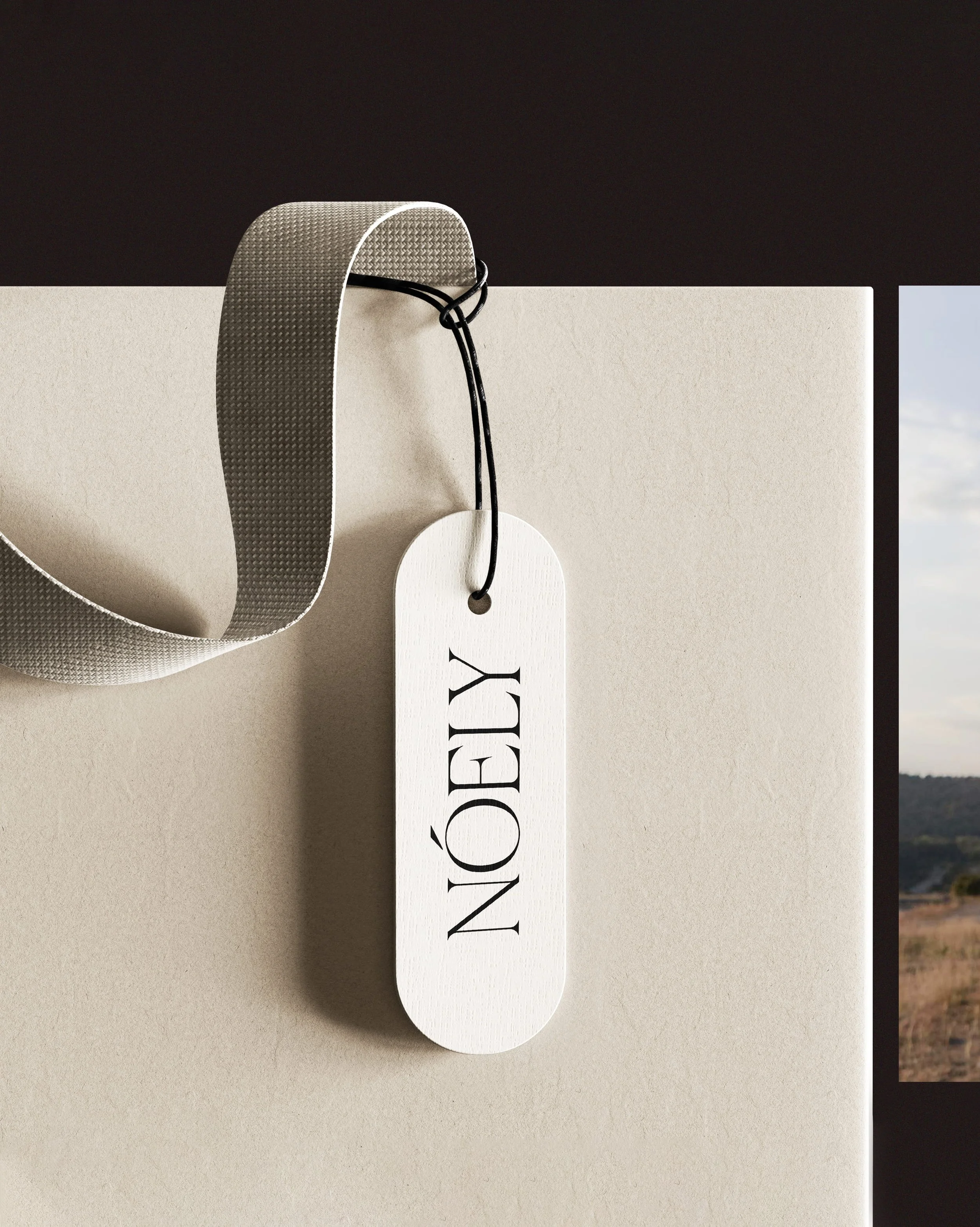 Close-up of a beige box with a white tag that has the brand name 'NOÉDC' in black letters hanging from a black string, with a grey textured strap looping over the box, against a dark background.