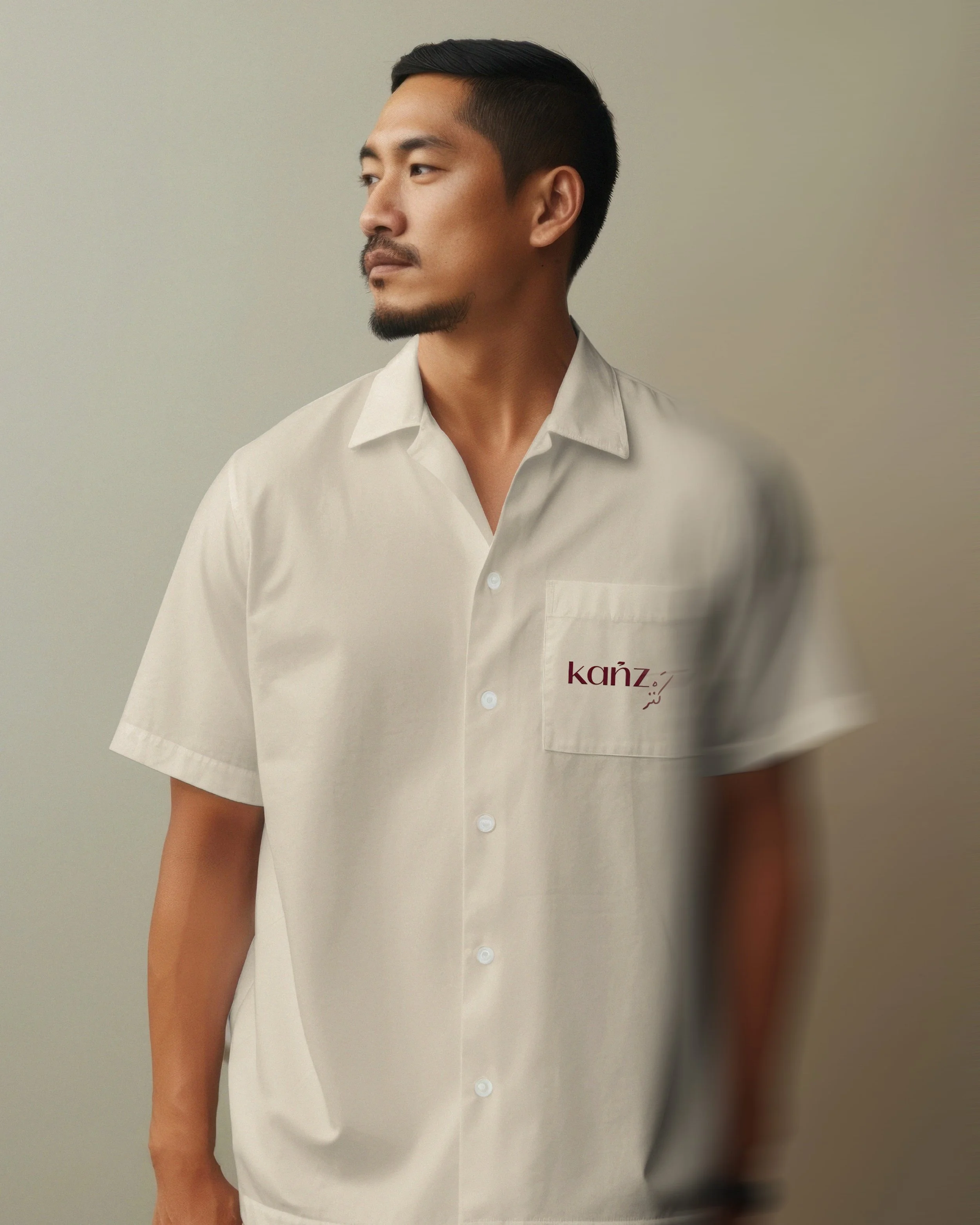 A man wearing a white shirt embroidered with the word 'Kanz' in red, standing against a plain background, looking to his left.