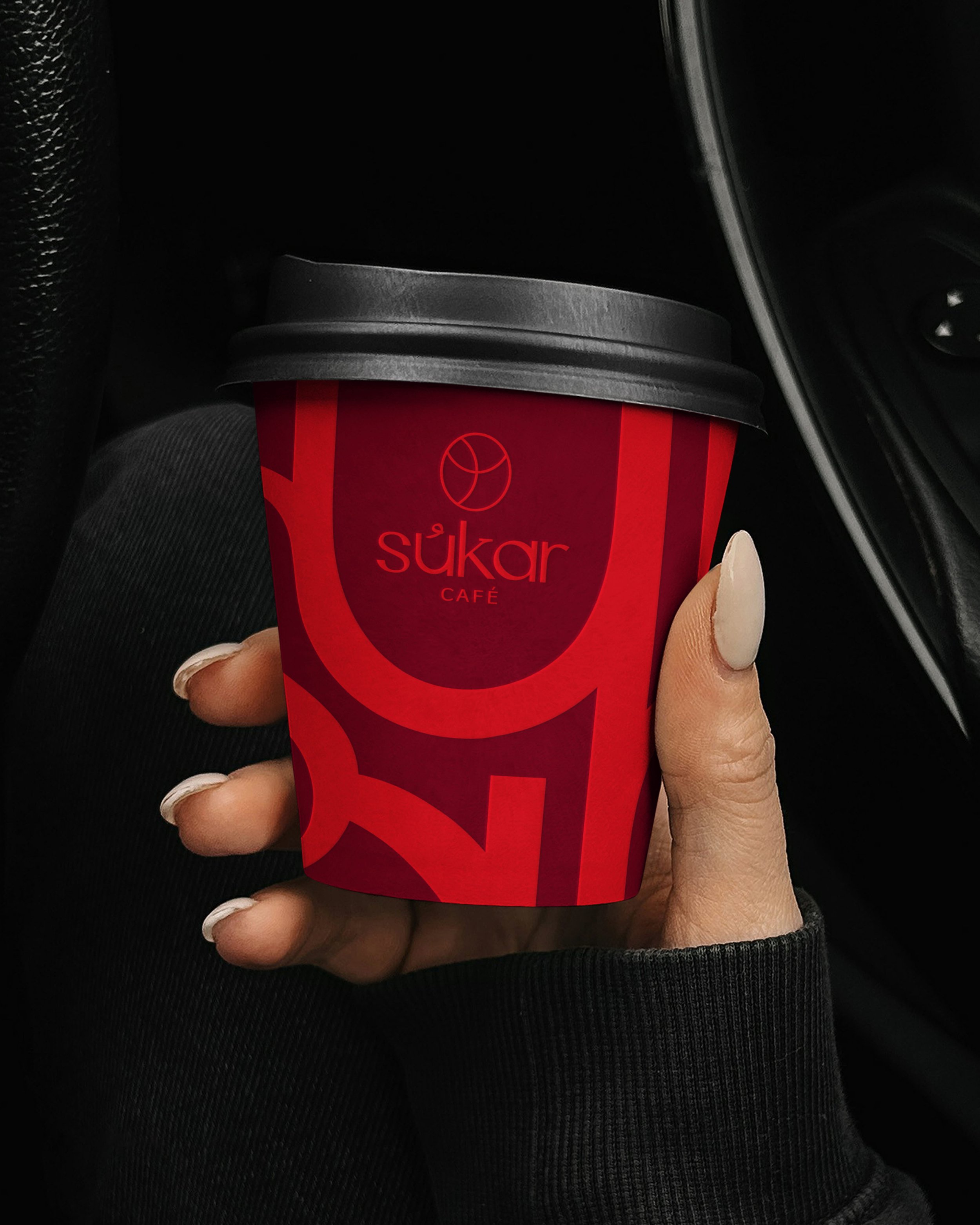 Close-up of a hand holding a red and black paper coffee cup with the words 'sükar café' and a logo, in a car interior.