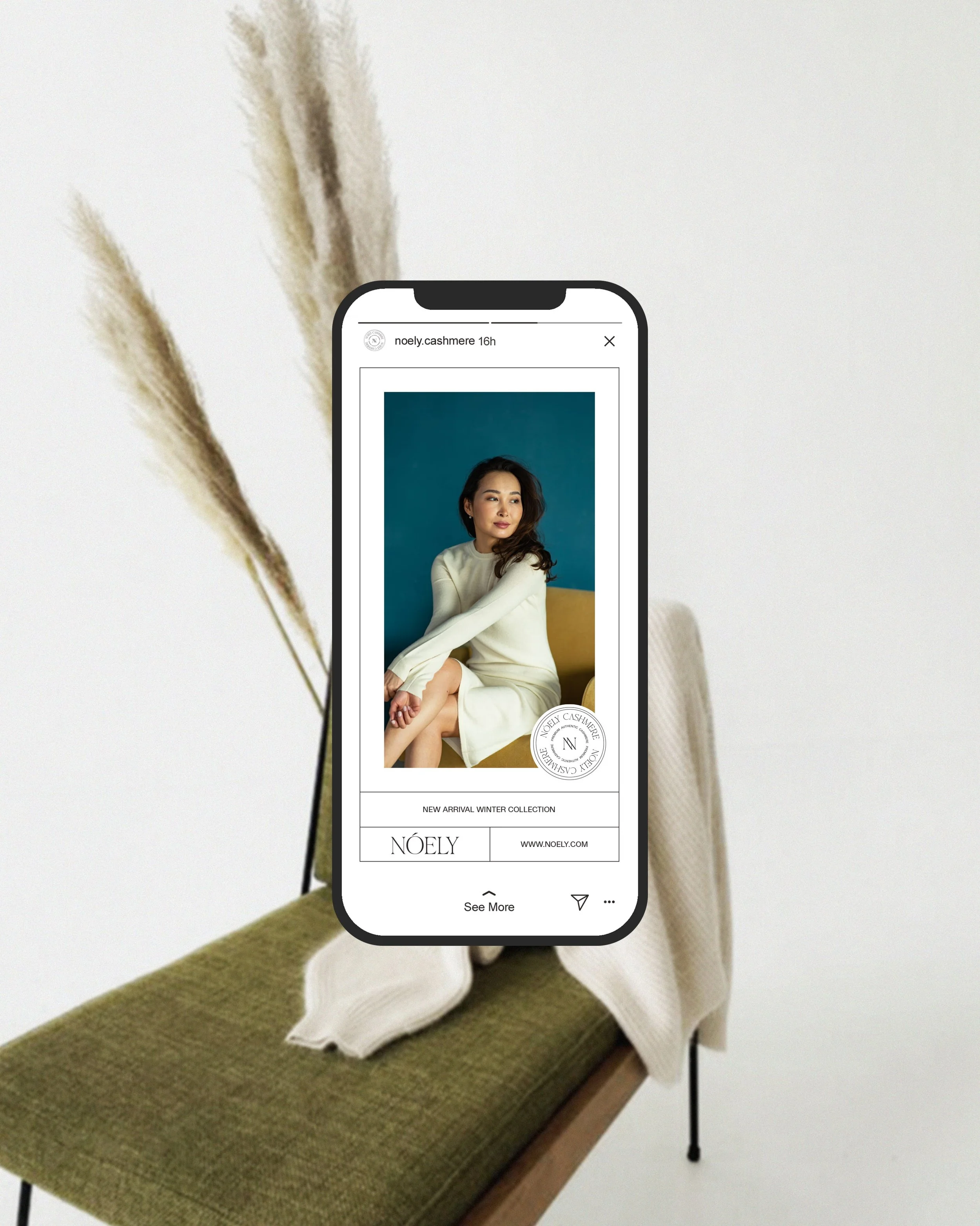 A smartphone shows a fashion advertisement for Noely Cashmere's winter collection, featuring a woman with dark hair, wearing a cream-colored dress, sitting against a teal background. The phone is held over a green cushioned bench with a cream-colored