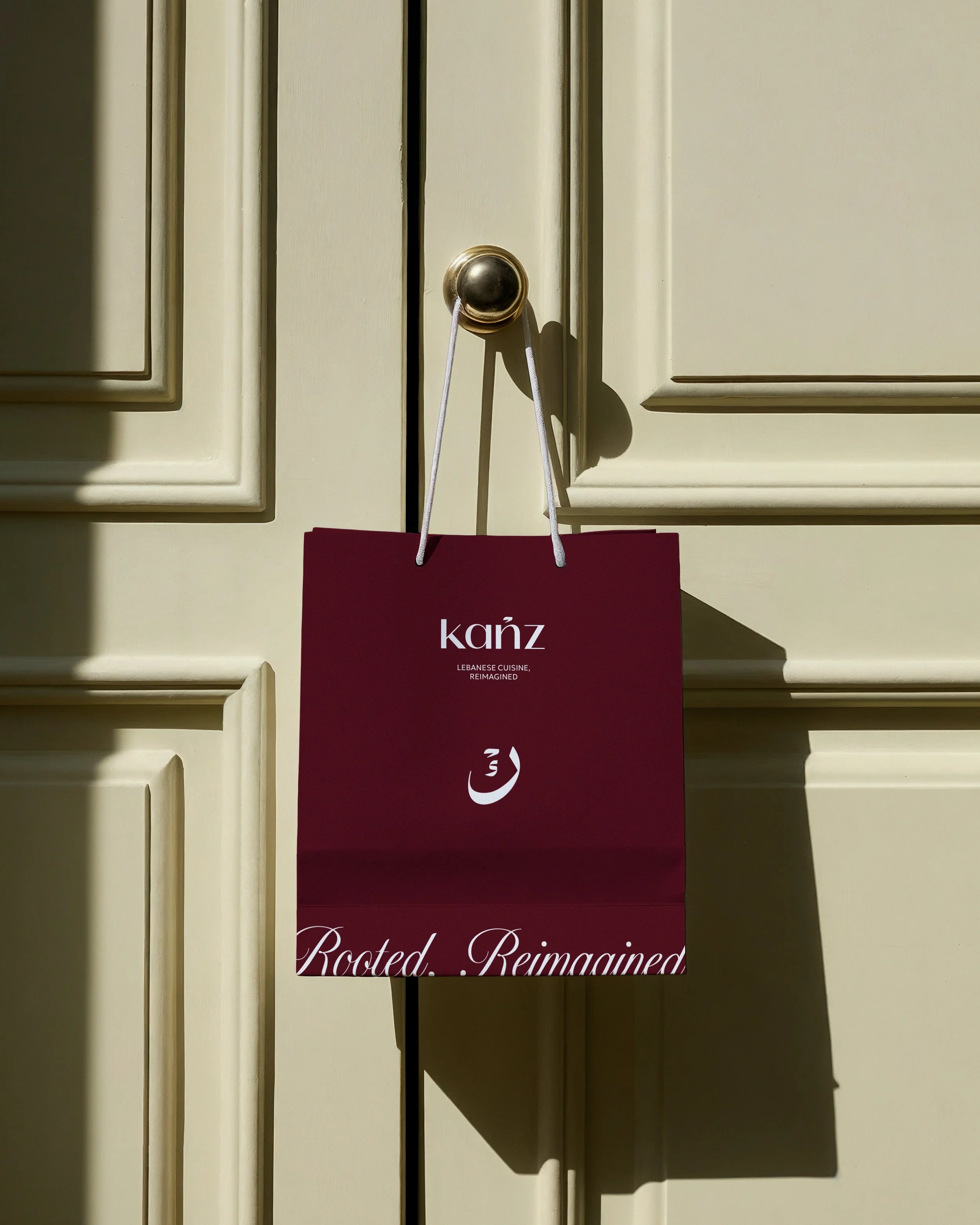 A maroon shopping bag hanging from a door handle with the text "Kanž" and below it "Lebanese Cuisine, Reimagined" in smaller white letters.