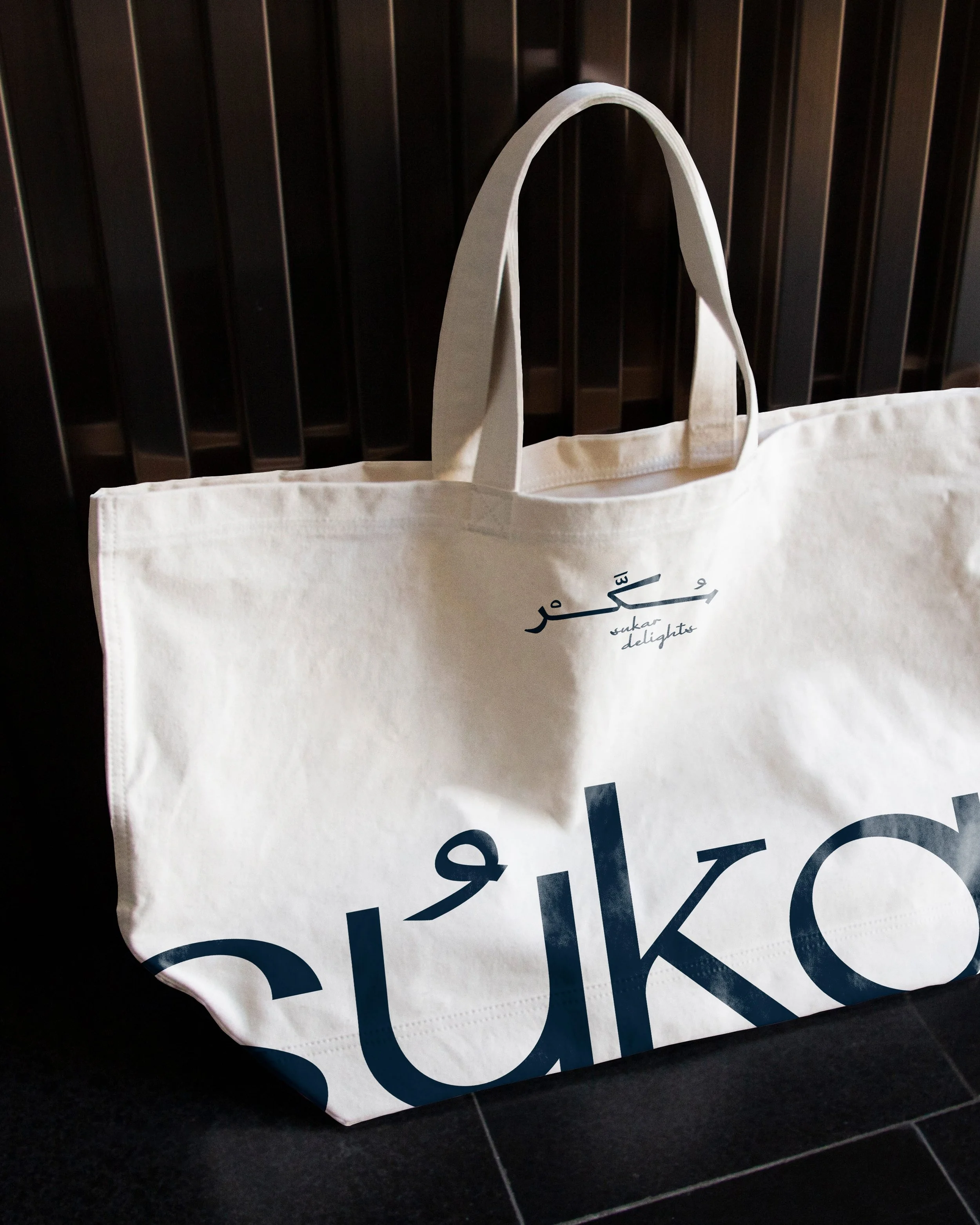 A white canvas shopping bag with dark blue text that reads 'sukkar delights' and some writing in a different script, placed on a black tiled floor in front of a black wooden slatted wall.