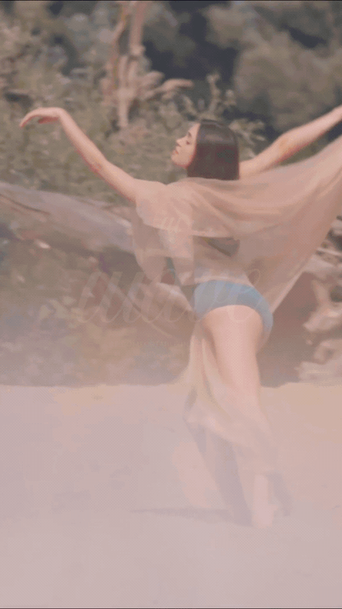 A woman standing outdoors on a beach, dancing with her arms extended, wearing a sheer cover-up and blue shorts.
