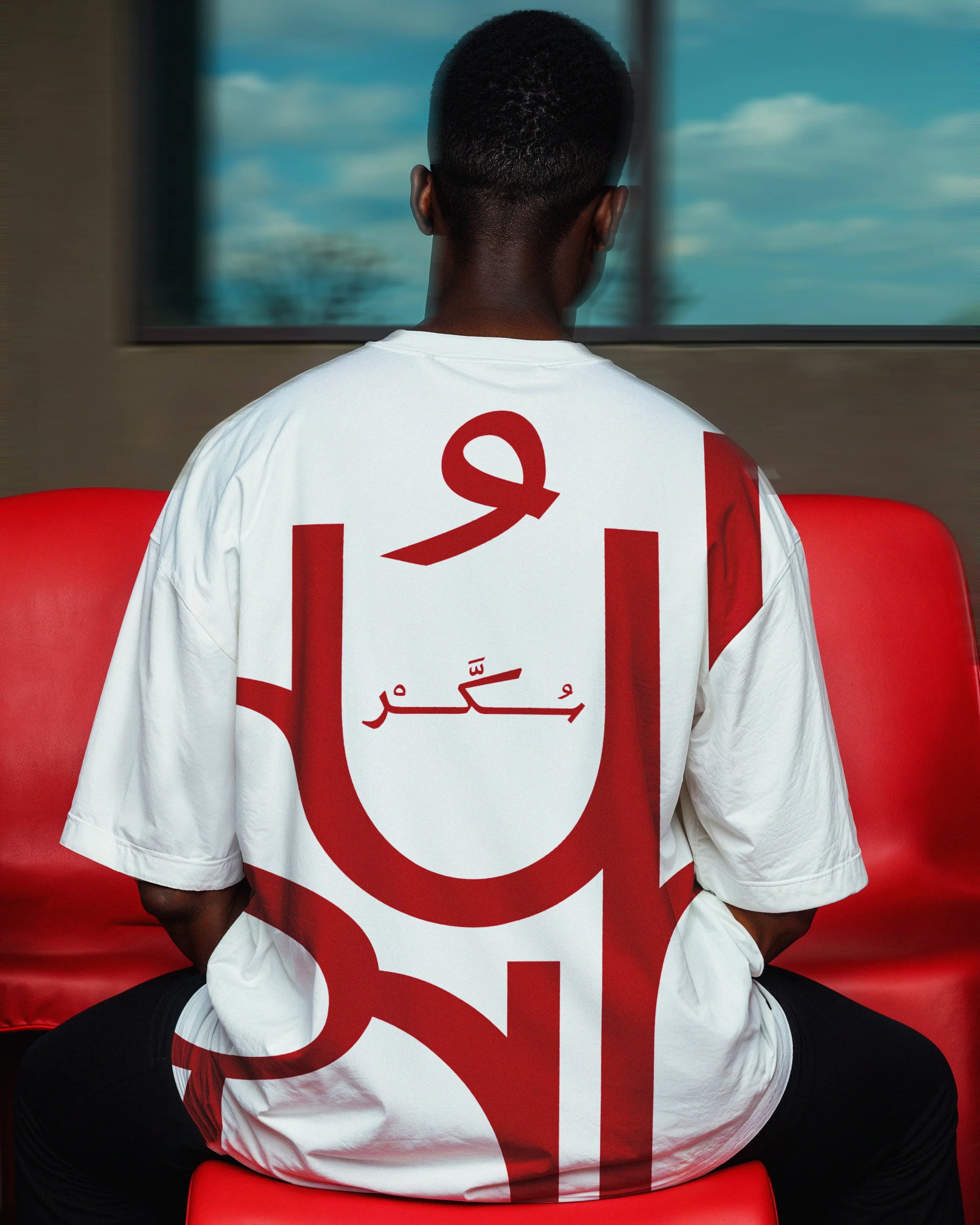 Back of a person sitting on a red chair, wearing a white T-shirt with red Arabic script and large red style ampersand on the back, facing a window with a view of a blue sky with white clouds.
