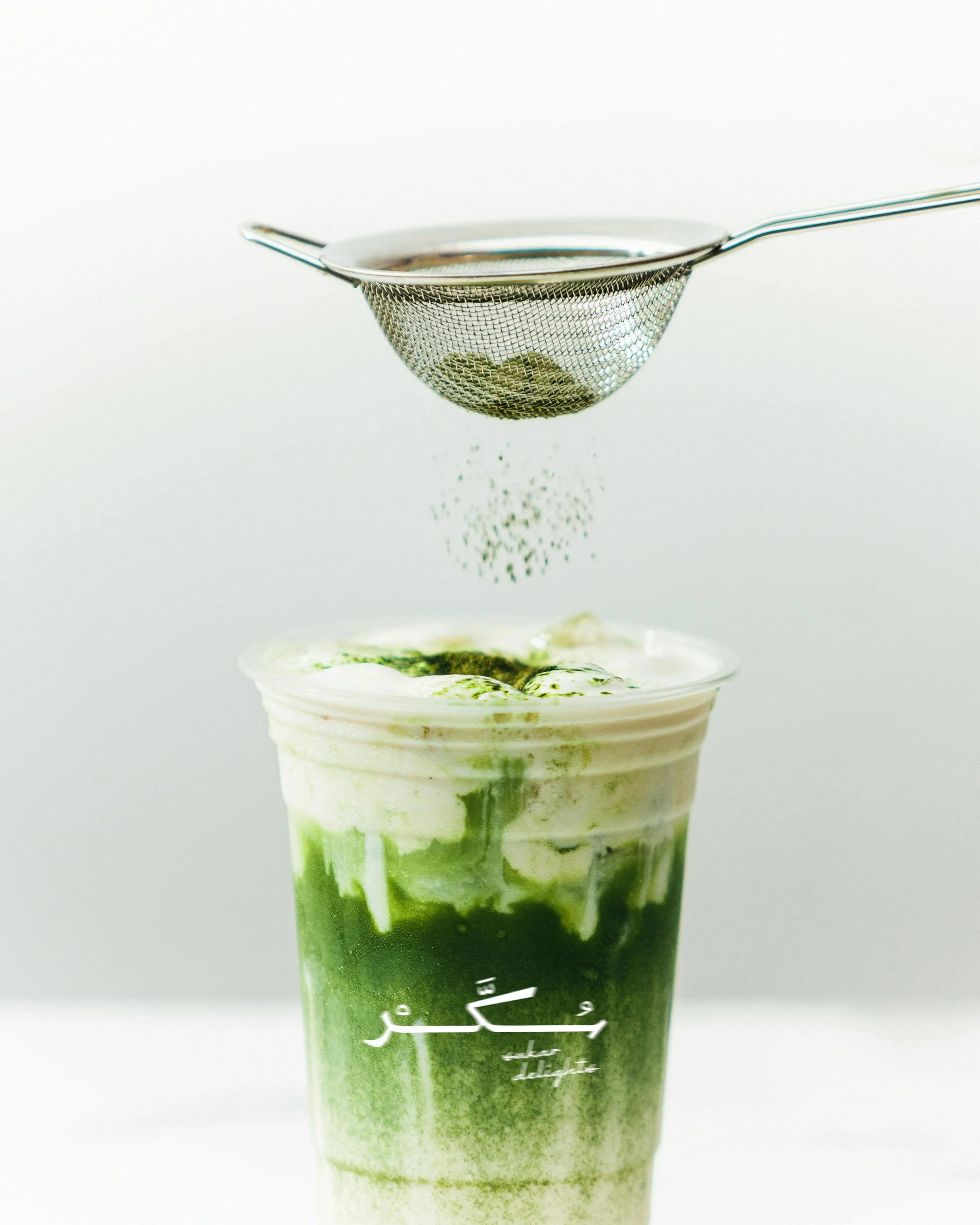 Green layered drink in a clear plastic cup with a white whipped topping and green sauce, with a metal sieve strainer above it, against a white background.