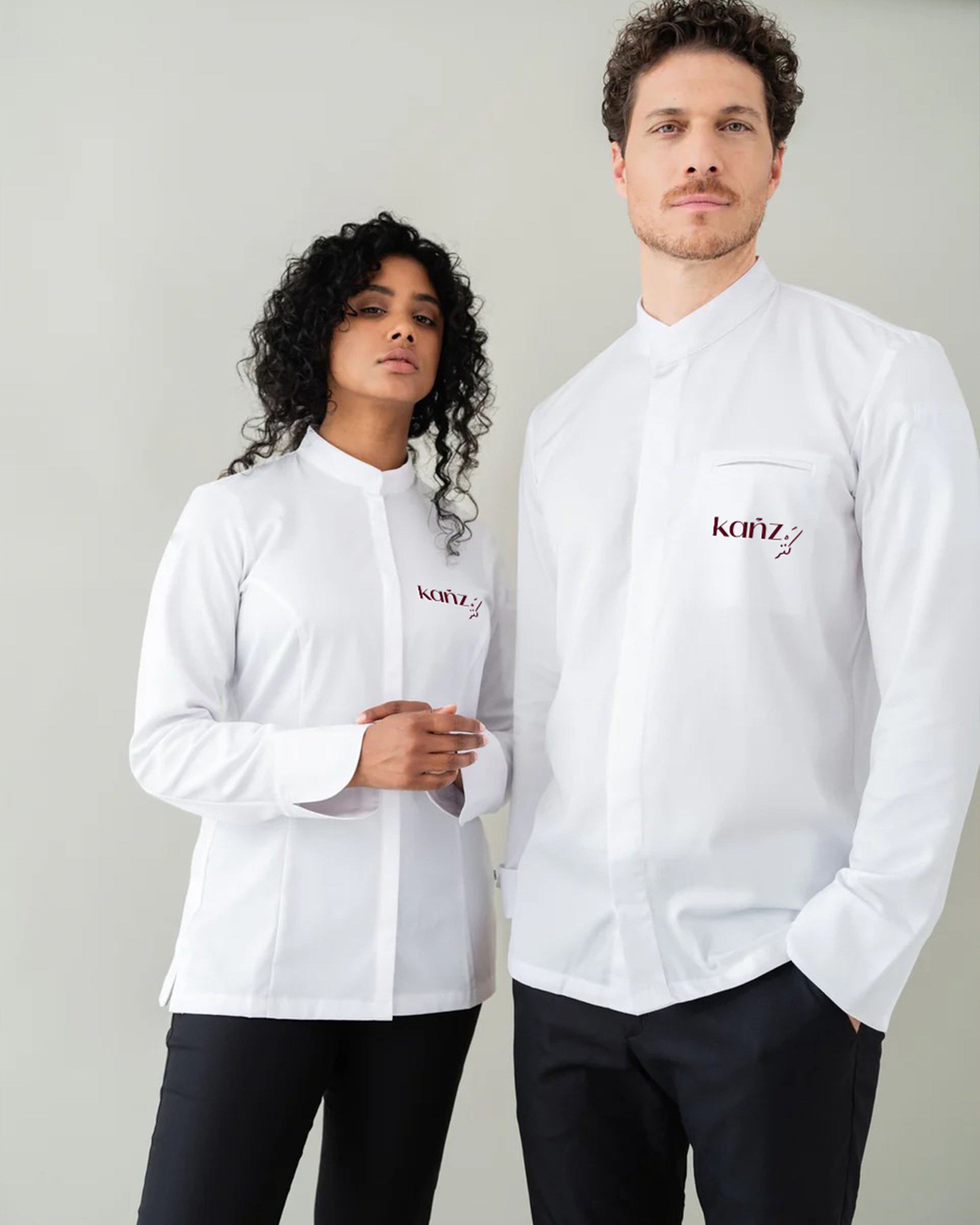 Two models standing side by side wearing white chef jackets with the logo "Känz" on the chest. The background is plain and neutral. The female model has curly dark hair and is looking at the camera with a neutral expression. The male model has short 