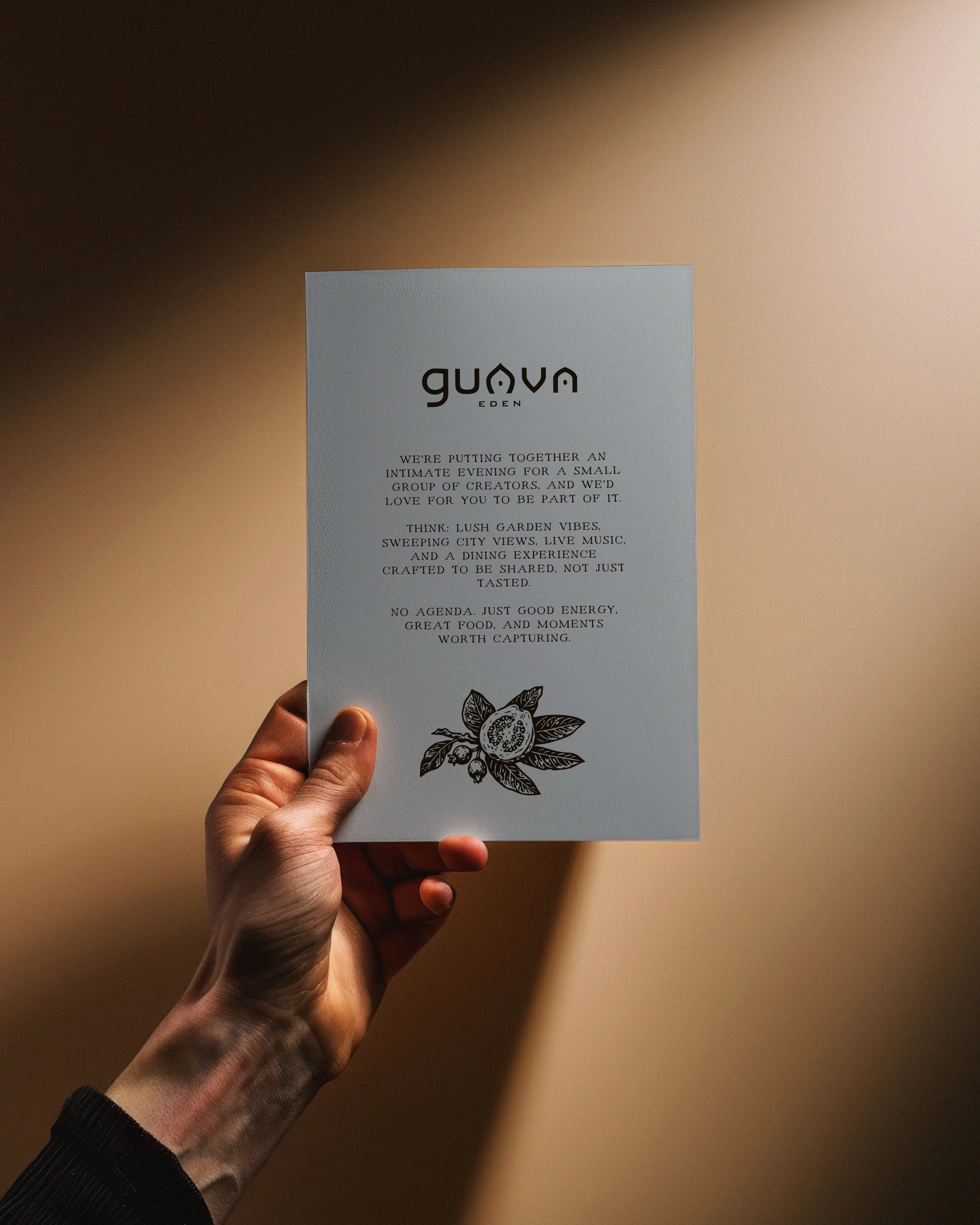 Hand holding a flyer with The Guava Eden logo and text about an intimate evening event with garden vibes, live music, and shared dining experience, featuring a black and white illustration of a guava flower.