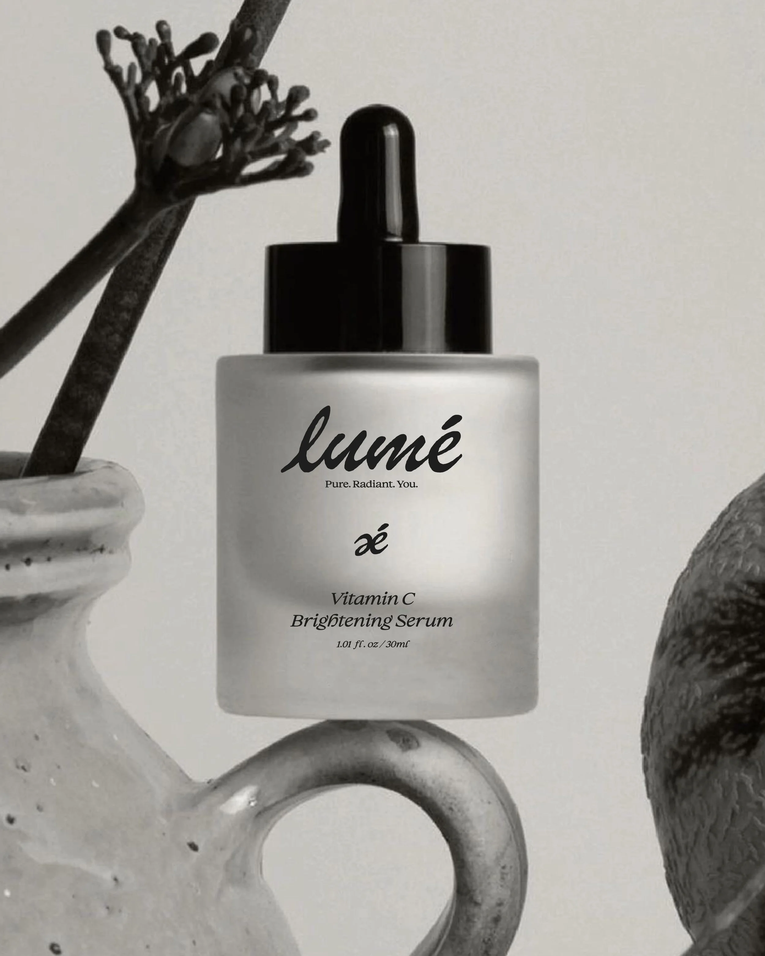 A white bottle labeled 'lumé' with the words 'Pure. Radiant. You.' and 'Vitamin C Brightening Serum' is centered next to a black dropper cap. To its left, there is a small marble container holding a dried flower or seed pod, and to the right, a round