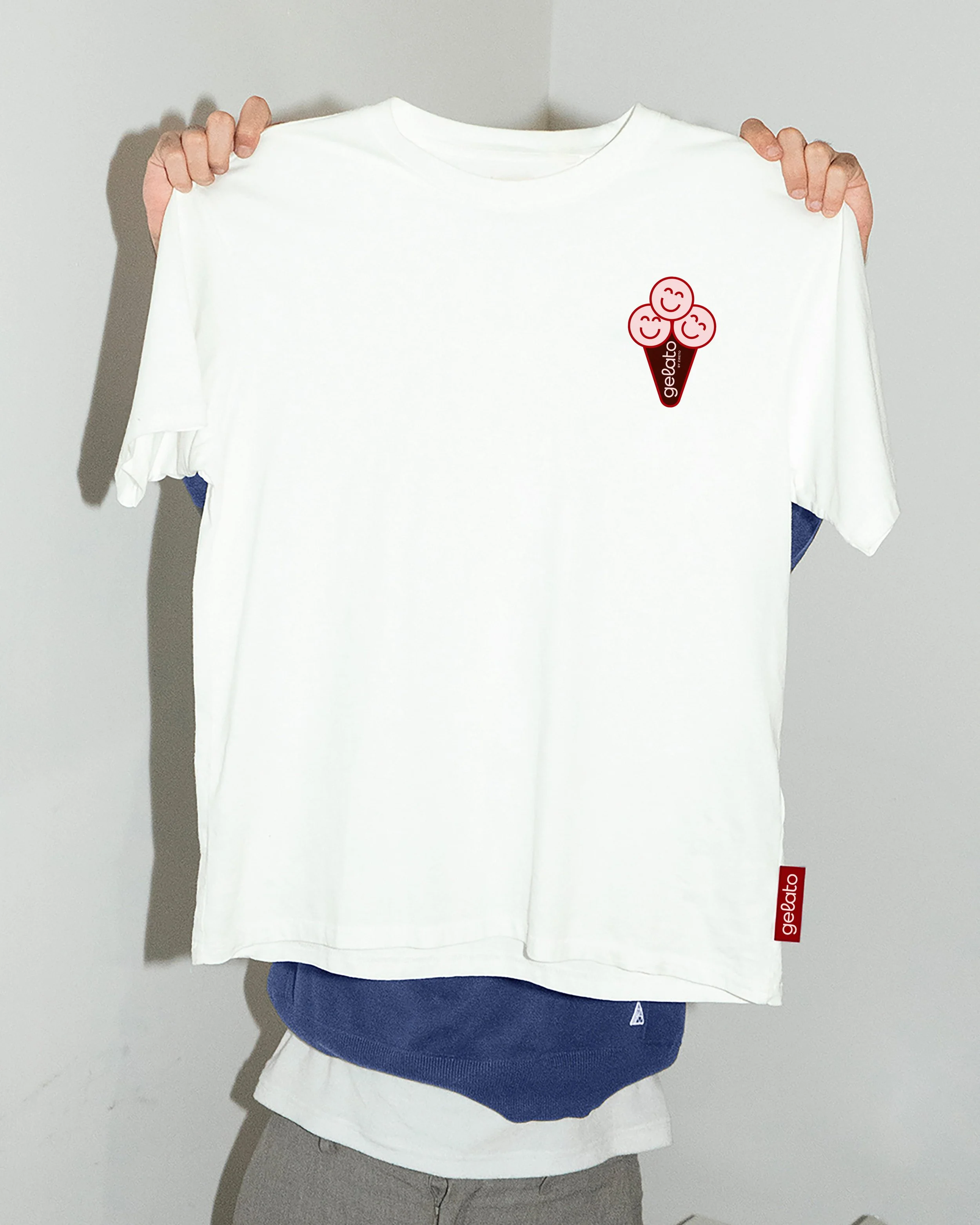 White T-shirt with a small logo on the left chest area showing three smiling faces inside an ice cream cone with the word 'Gelato' beneath.
