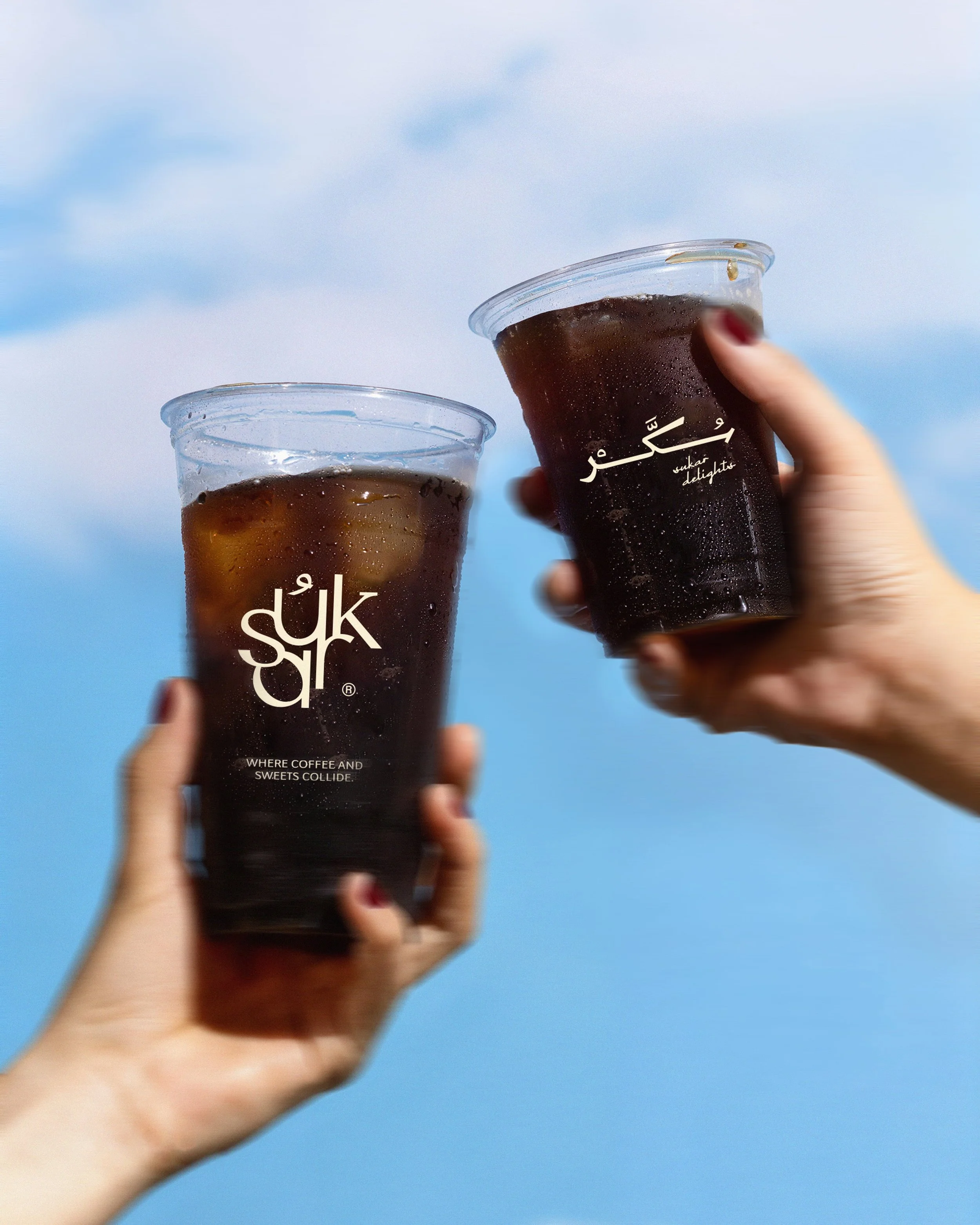 Two hands holding cups of iced coffee against a blue sky background.