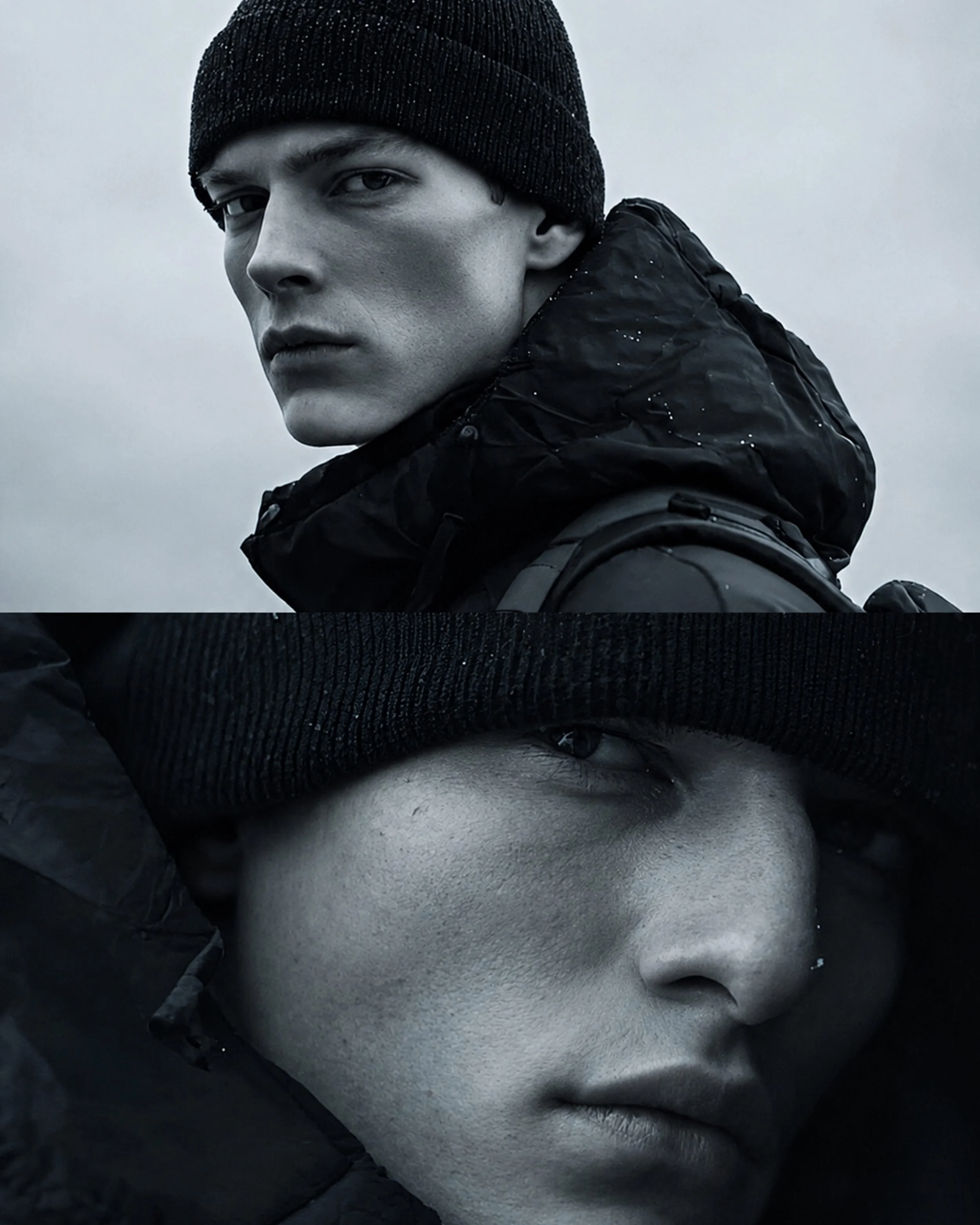 A black and white photo of a young man with short hair, wearing a black beanie and a dark jacket, looking over his shoulder with a serious expression, against a cloudy sky background.
