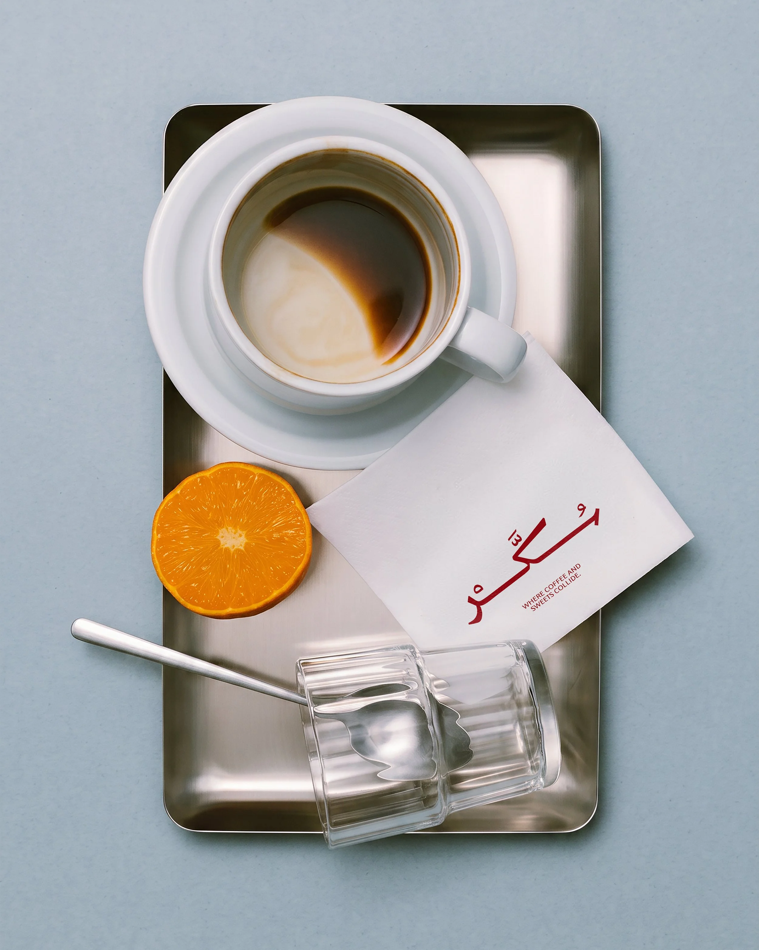 Top view of a metal tray with a white cup of black coffee, two orange slices, a white paper with red text, and two transparent plastic cups.
