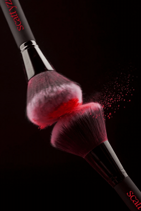 Two makeup brushes, one releasing red powder onto the other, against a black background.