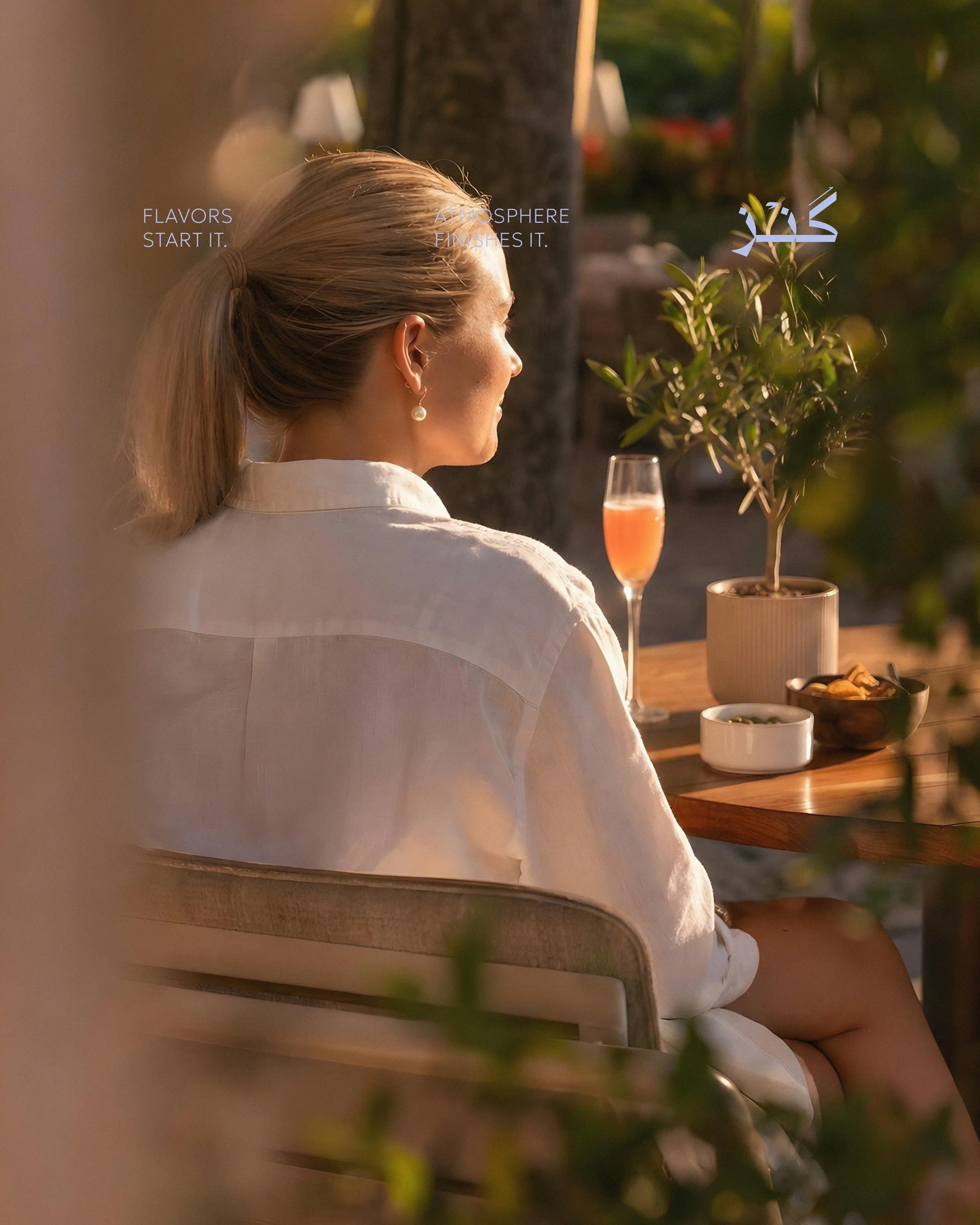 A woman with blonde hair tied back in a ponytail, wearing a white shirt, sitting at an outdoor table with a glass of pink beverage, potted plant, and bowls of food, illuminated by warm sunlight with a blurred background of trees and outdoor furniture