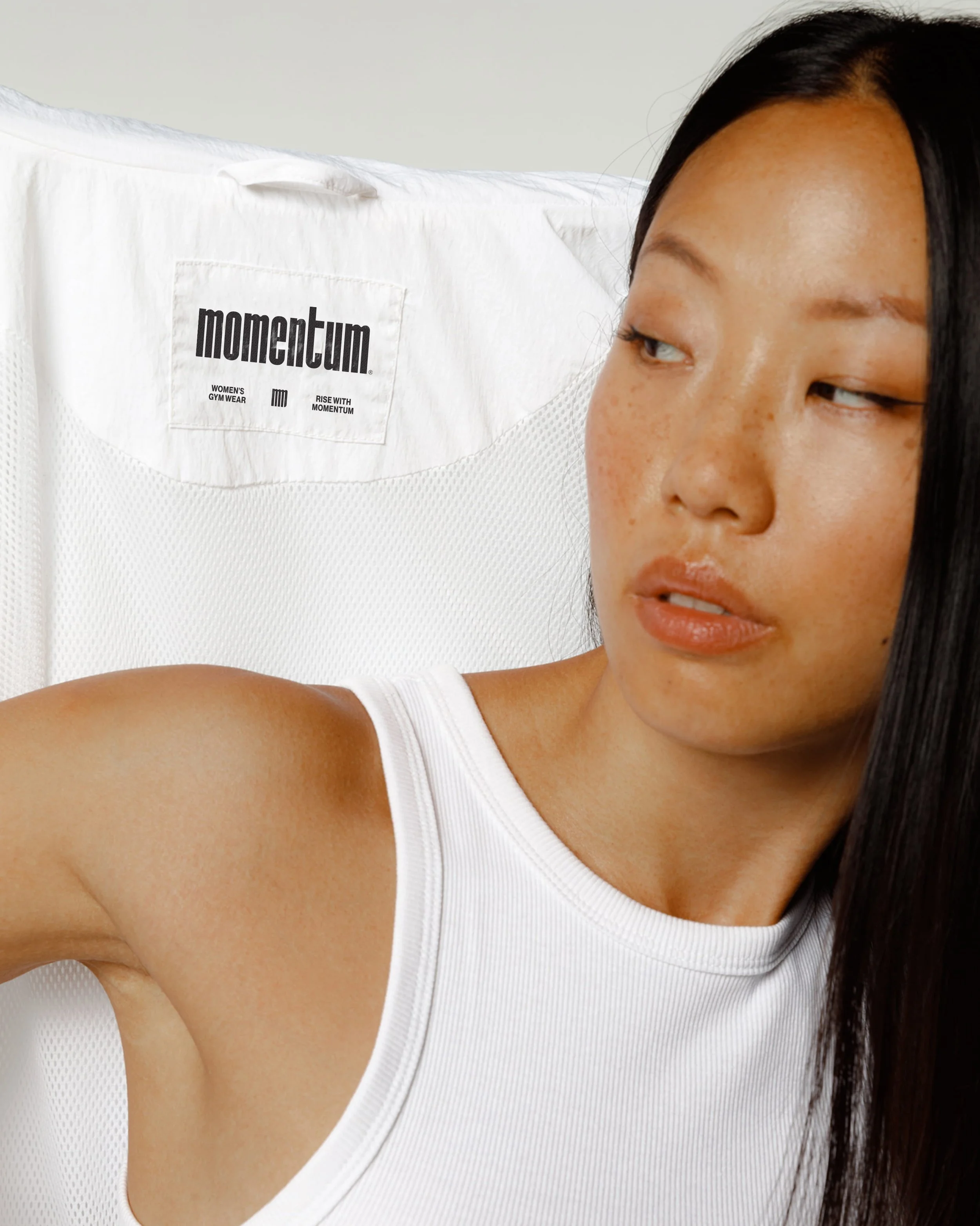 Close-up of a woman with dark hair wearing a white sleeveless top, taking a selfie in front of a white background with a visible tag that reads 'momentum Women's Gym Wear Rise with Momentum'.
