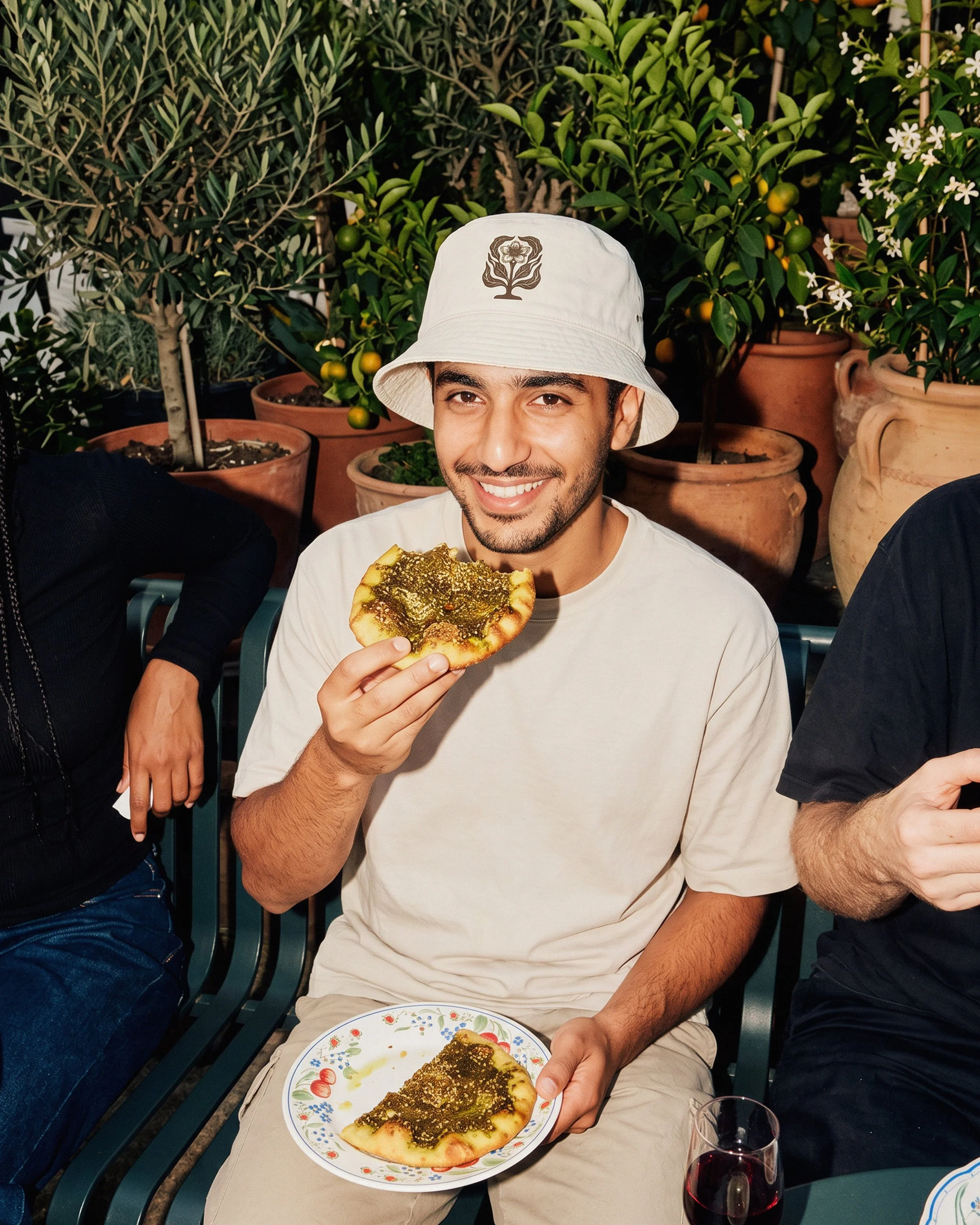 A man wearing a white bucket hat with a floral design smiles while holding a slice of pizza topped with green pesto and cheese. He is seated at a table with a porcelain plate containing the same pizza slice. Behind him, there are potted orange trees 