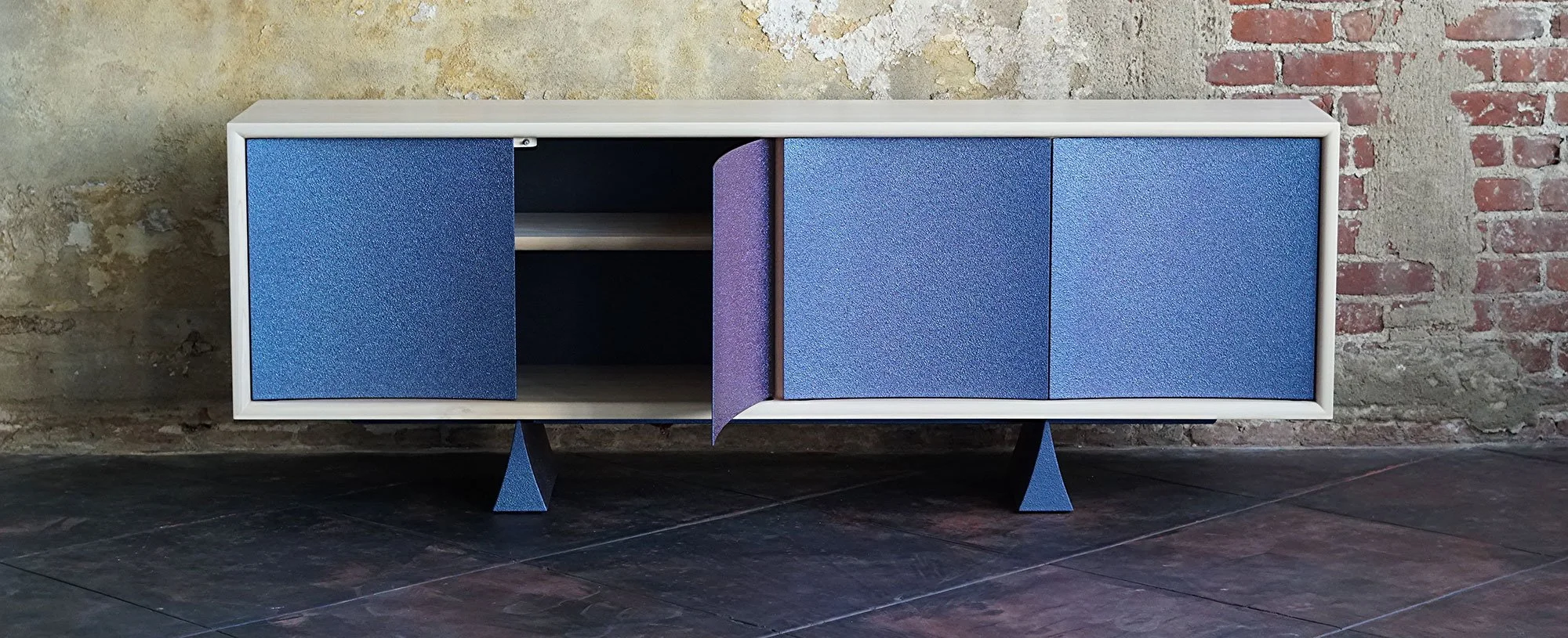 Modern wooden cabinet with three doors, two blue and one purple, on a black stand with triangular legs, set against a textured brick and concrete wall.