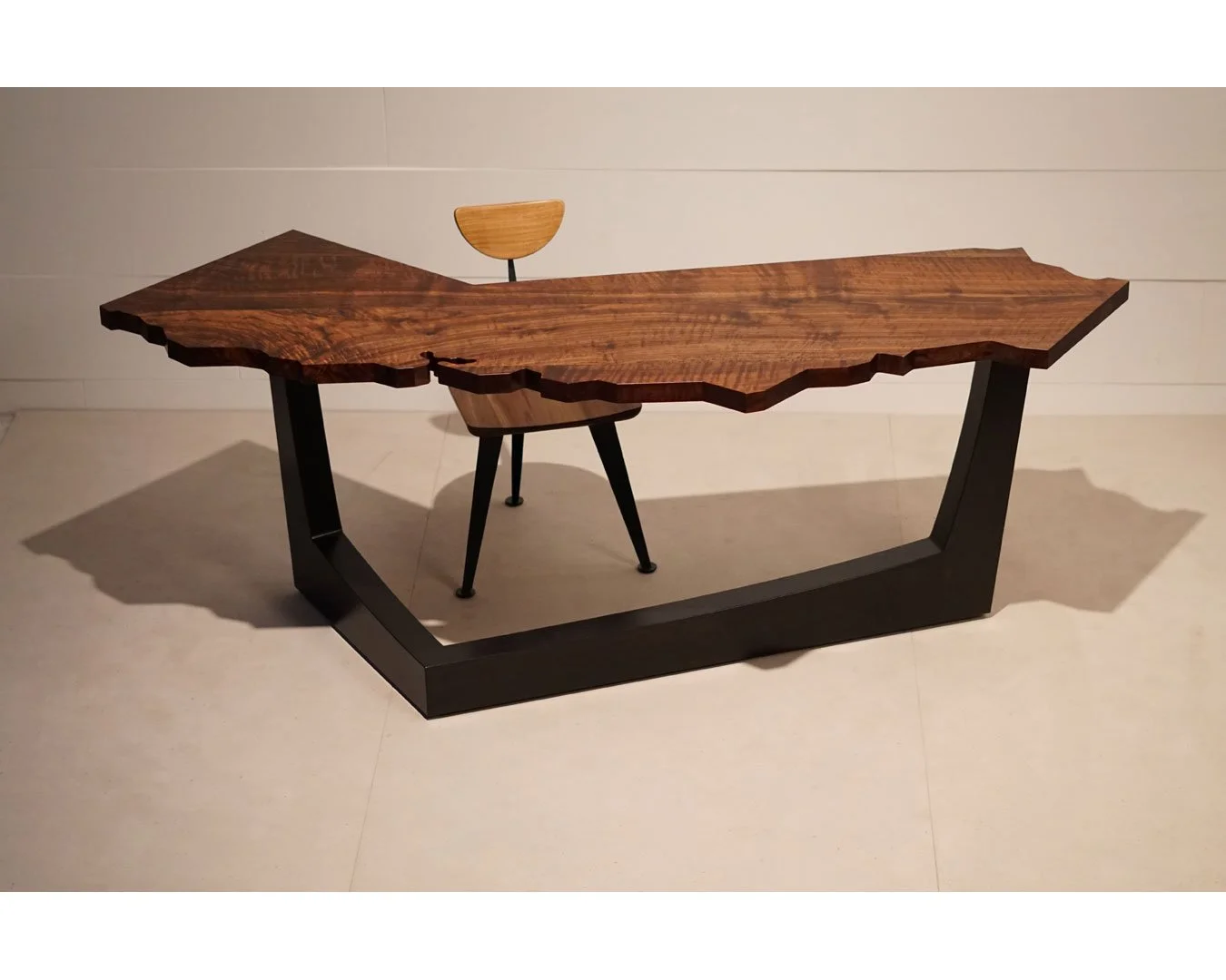 A modern wooden table with a irregular, natural edge top, supported by a black geometric metal base. There is a small, curved wooden chair in the background, placed behind the table.