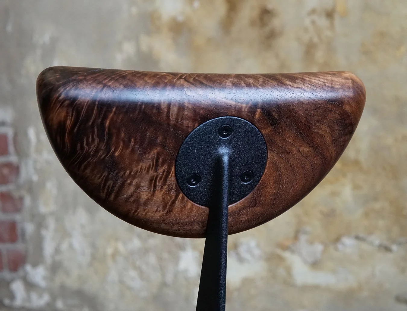 Close-up of a wooden object with a black mounting plate and a black arm attached, set against a mottled beige and gray background.