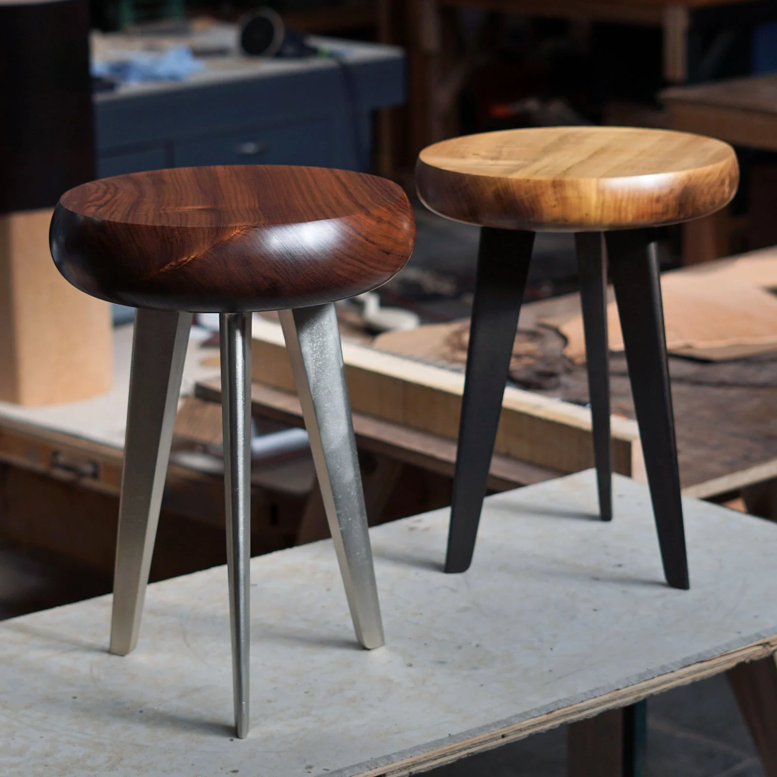 Two wooden stools with three-legged metal bases, one with a darker wood top and the other with a lighter wood top, placed on a worktable in a woodworking studio.