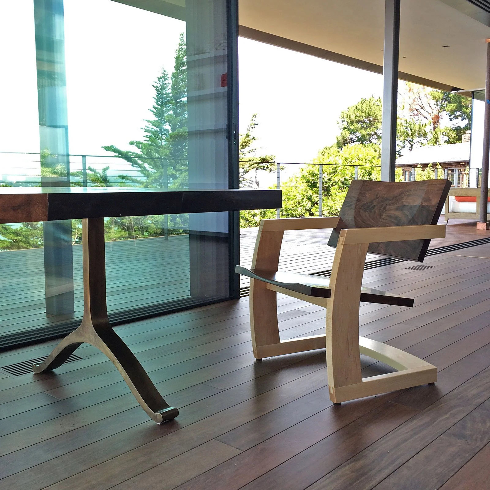 A modern interior space with large windows revealing outdoor trees, featuring a black wooden desk with curved legs and a minimalist wooden chair with a slanted dark wood seat and backrest.