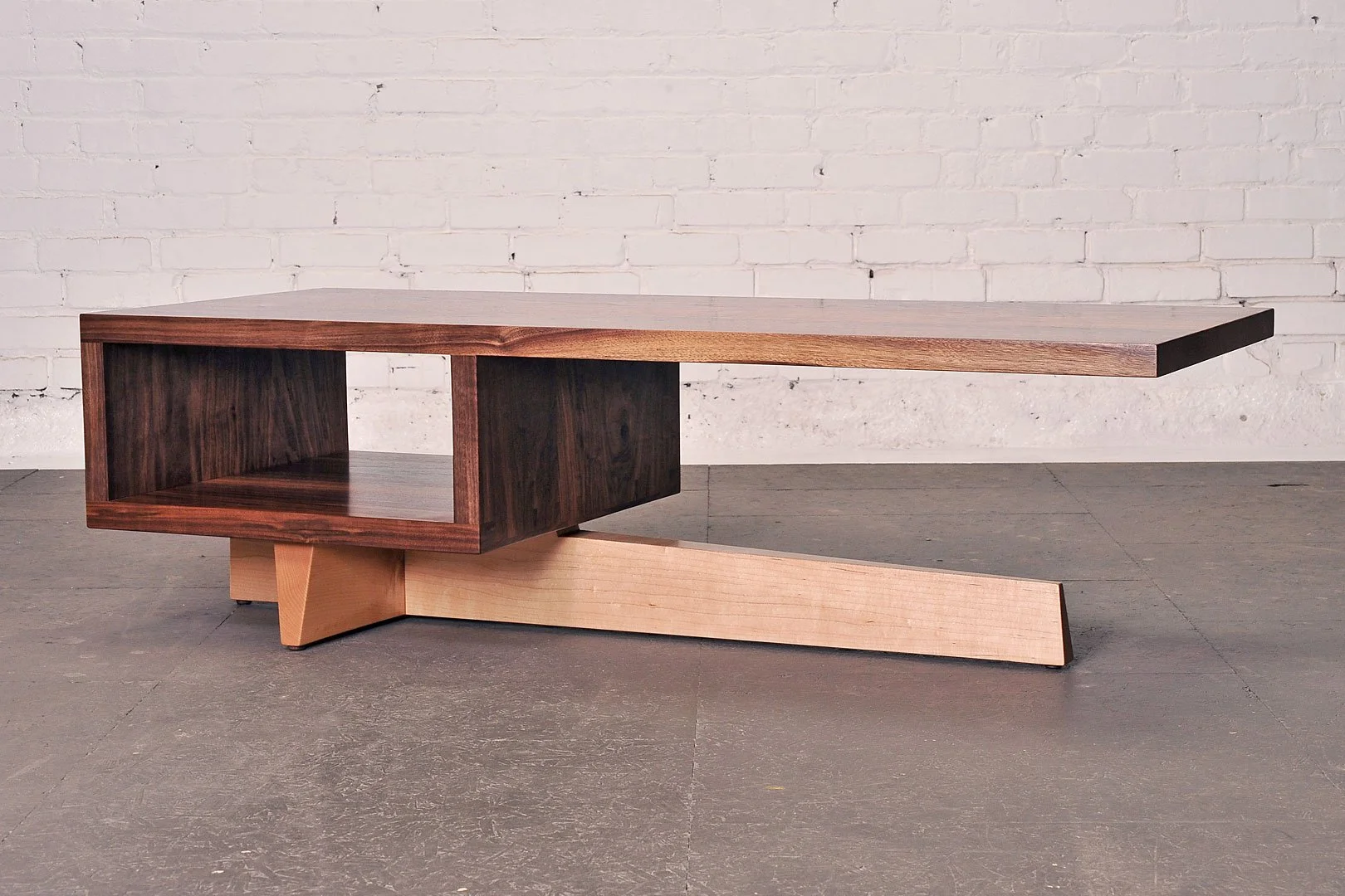 A modern, minimalist wooden coffee table with a rectangular open storage compartment on one side, set against a white brick wall and dark flooring.