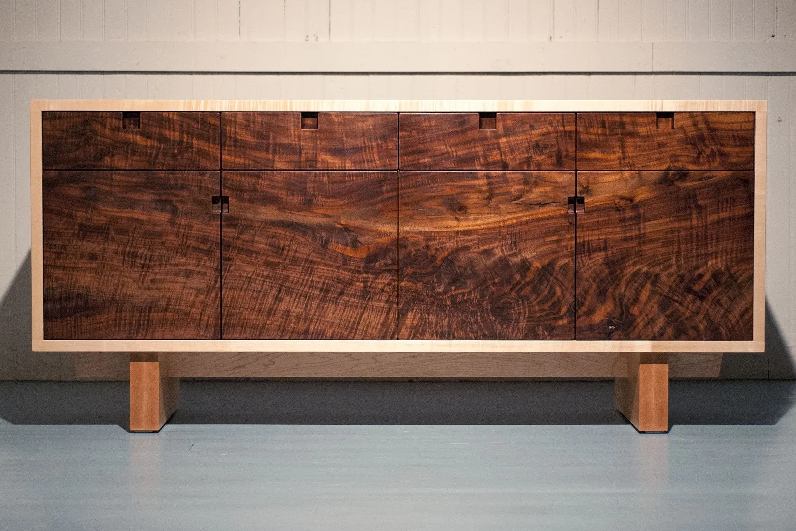 A wooden sideboard with a dark, patterned finish, standing on four light wooden legs, against a beige wall.