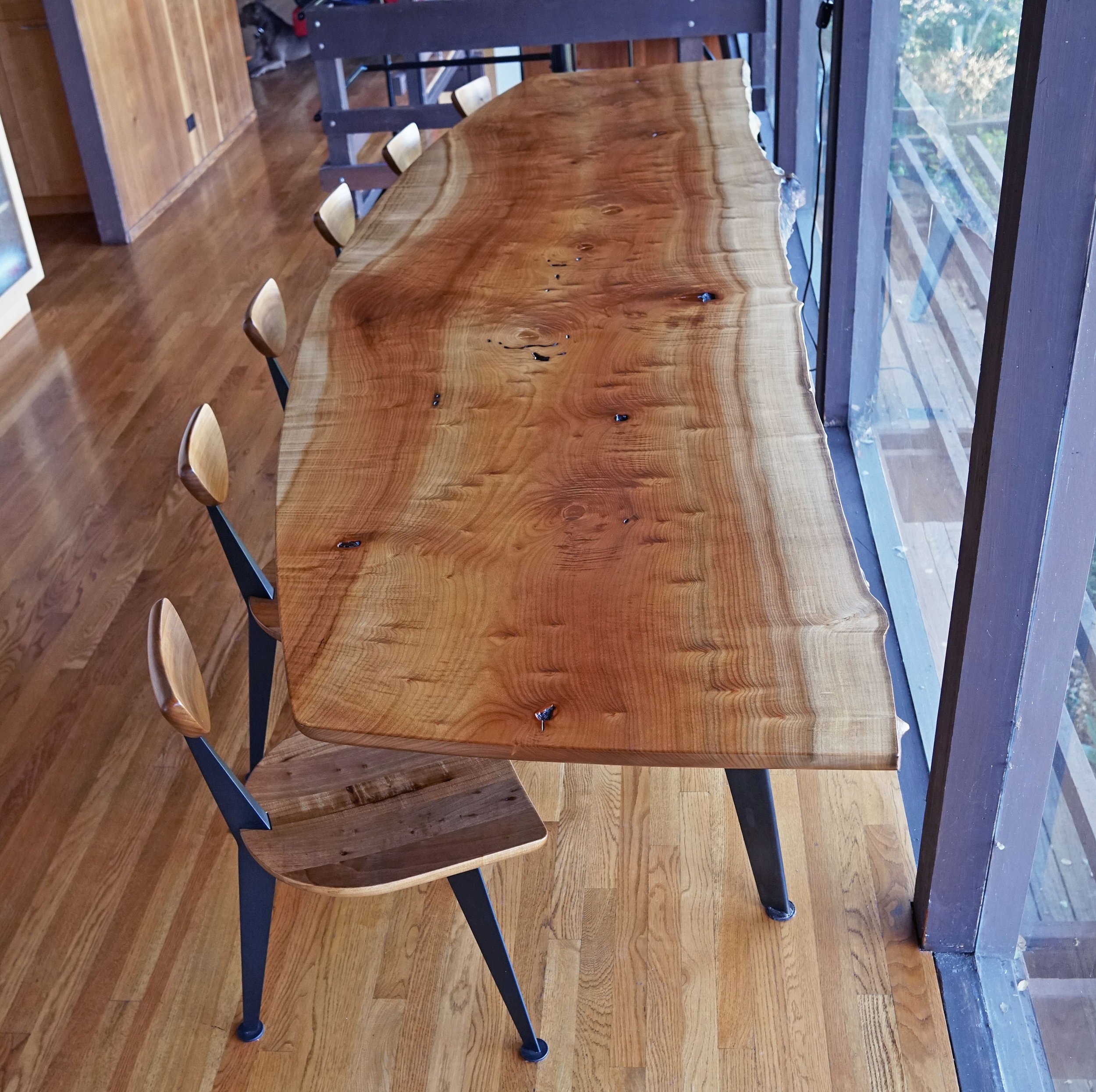 A long wooden dining table with live edge design in a room with hardwood floors, surrounded by wooden chairs, and a large window showing trees outside.