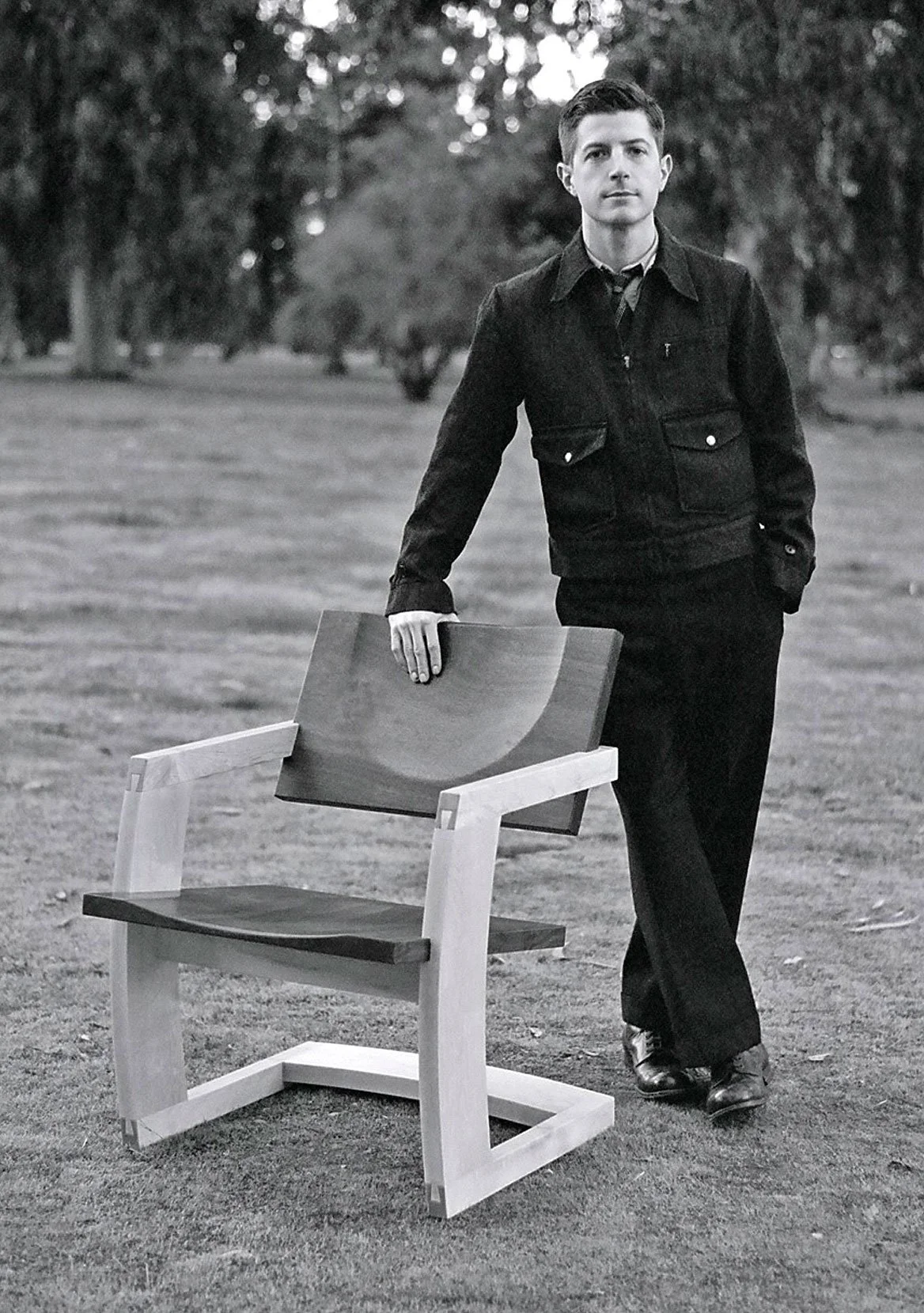 A young man in a black jacket and dark pants standing outdoors with one hand resting on a modern wooden and white chair on a grassy field, blurred trees in the background, in black and white.