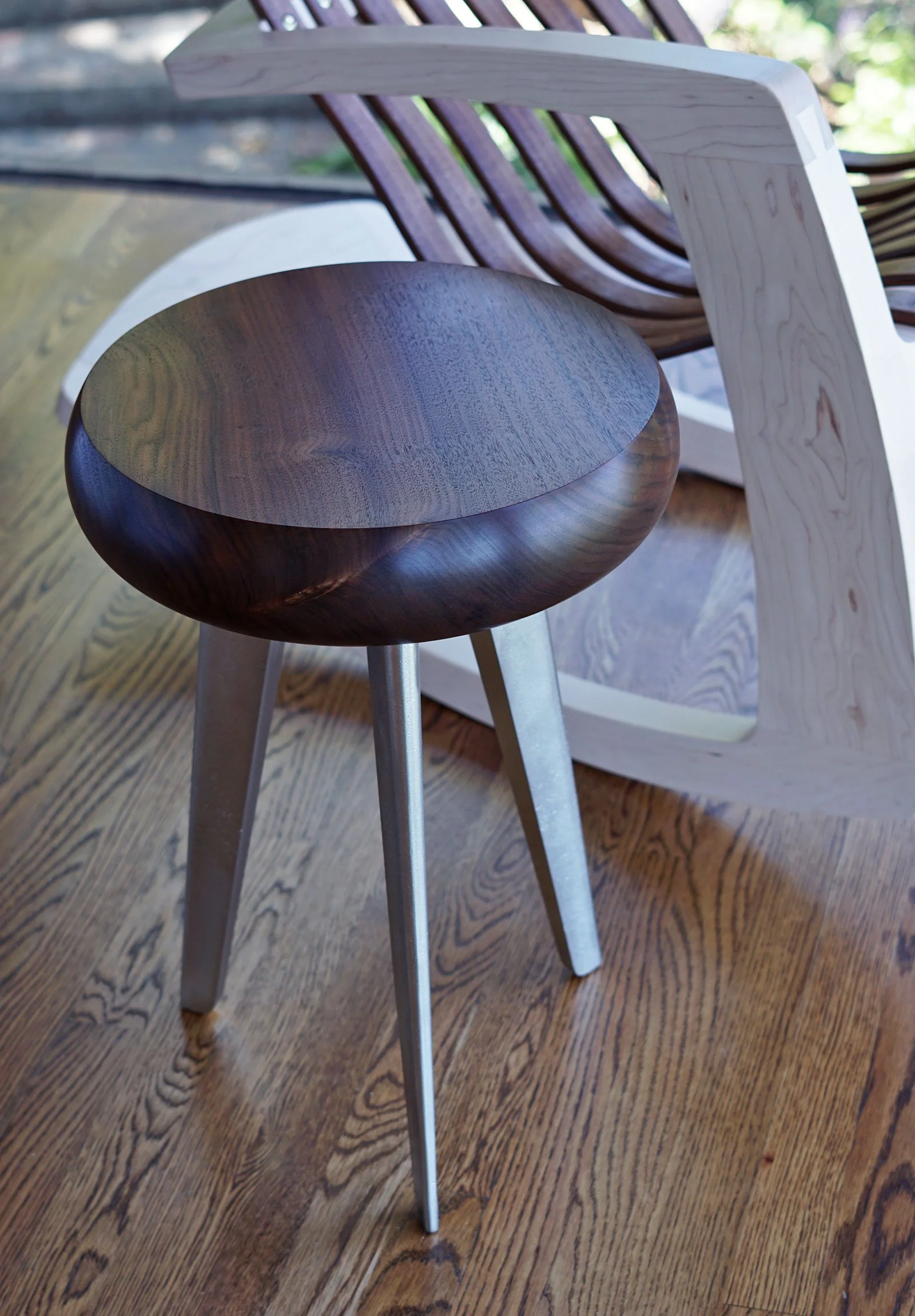 Close-up of a dark wooden stool with a rounded seat and light-colored metal legs, placed on a wooden floor. In the background, there is a wooden chair with curved backrest slats and a light-colored wooden armrest.