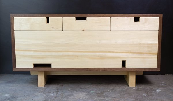 A wooden sideboard with a dark brown frame, light beige drawers, and a thick top, standing on four short legs against a dark wall.