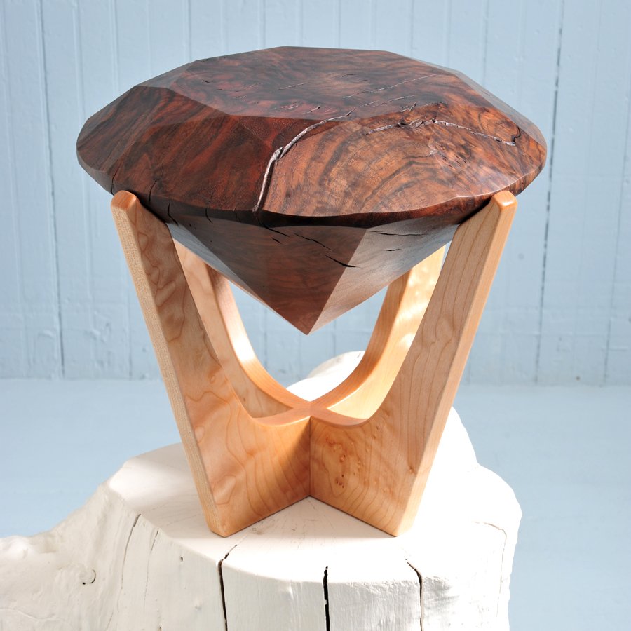 Wooden sculpture of a diamond shape supported by two light-colored wooden legs, with a dark, polished wooden top, placed on a white surface with a light blue background.