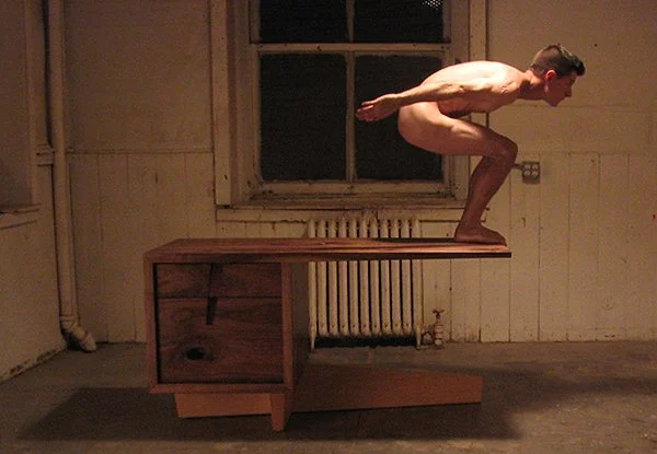 A man in a crouched position balancing on a wooden desk in a dimly lit room with a window, radiator, and wooden walls.
