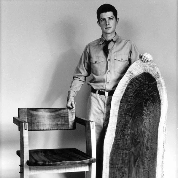 A young man in a button-up shirt, tie, and pants standing next to a wooden chair and holding a large, sliced piece of wood.