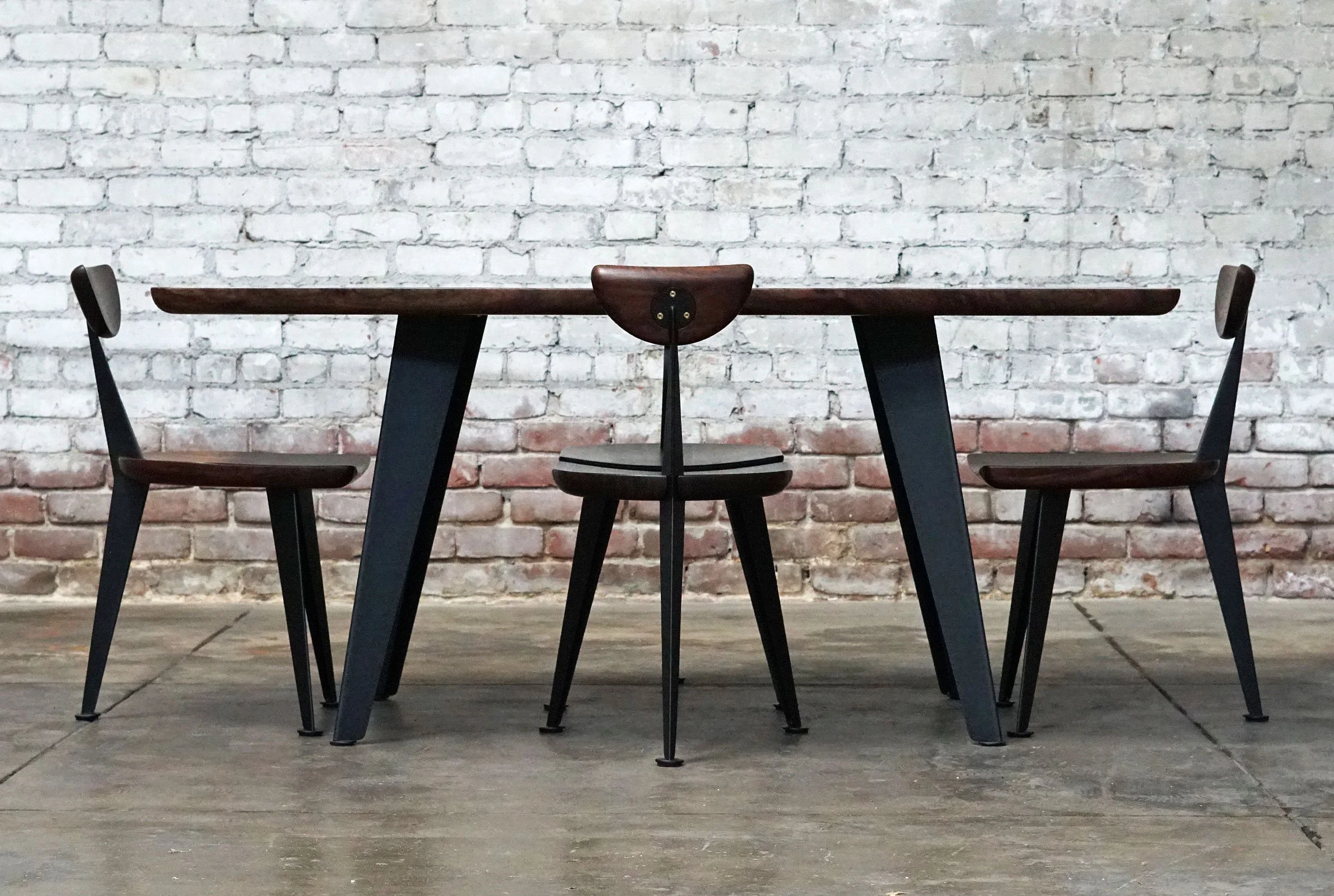 A wooden table with four black-legged chairs placed around it, set against a white brick wall in an industrial-style room with a concrete floor.