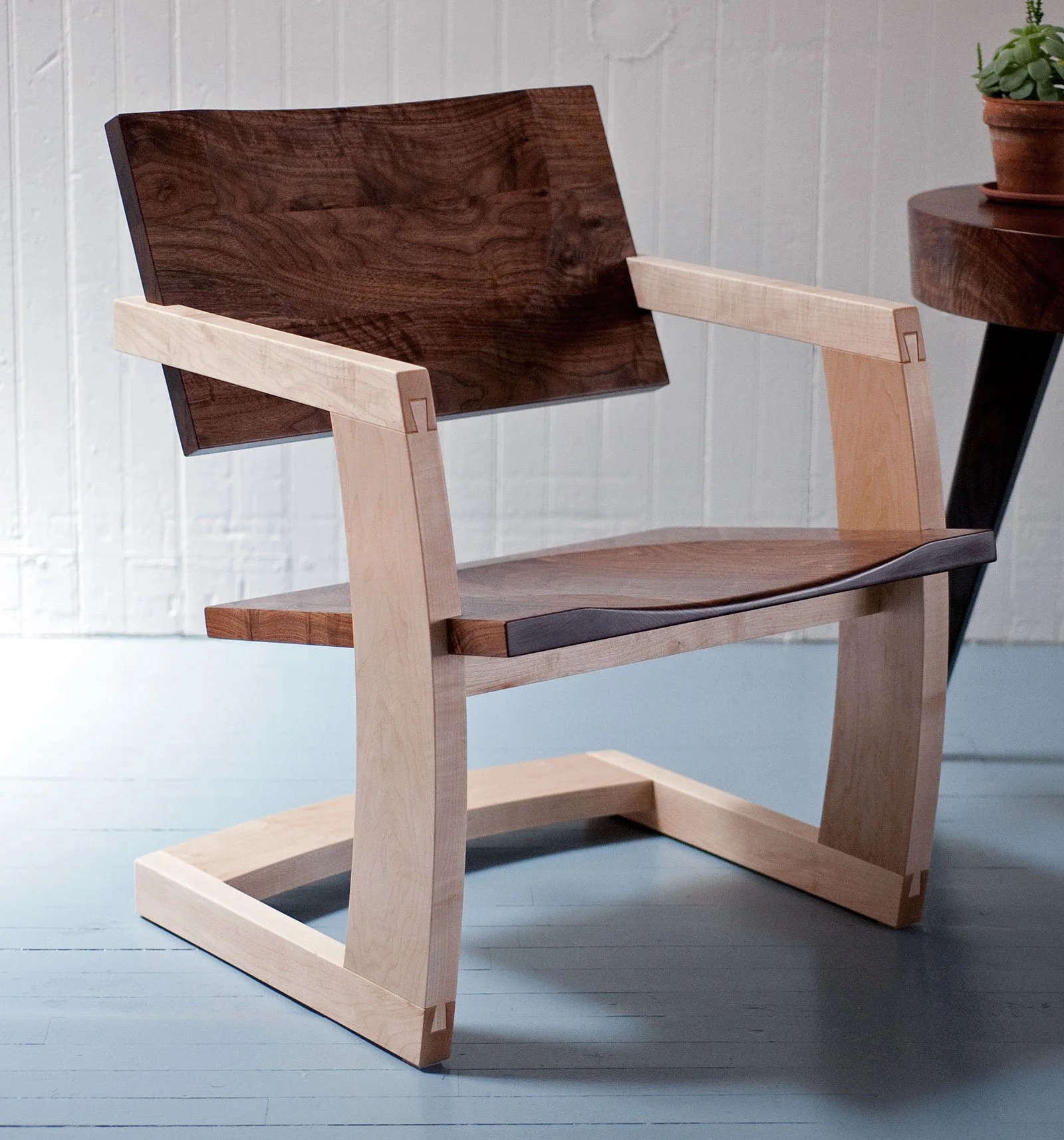 Modern wooden chair with a dark wood backrest and a lighter wood frame, positioned in a room with white paneled wall and light-colored floor.