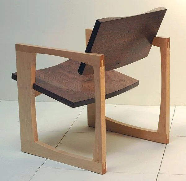 A modern wooden chair with a unique curved design, featuring a dark wooden seat and backrest, and light wooden armrests and frame.