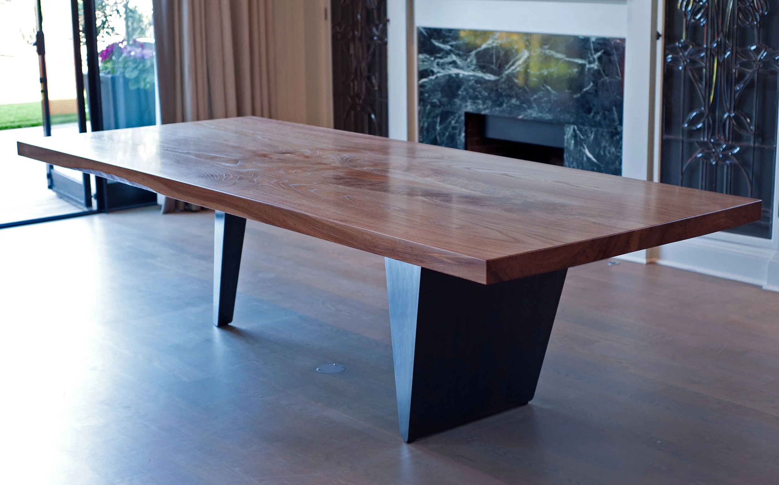 A wooden table with metal legs positioned in a modern living room, with a fireplace and decorative metalwork on the wall in the background.