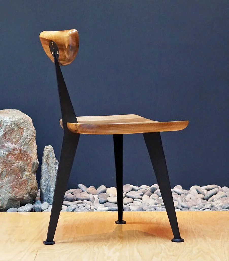 A modern wooden chair with black legs and a curved wooden backrest, positioned on a wooden surface against a dark blue wall with rocks and stones nearby.