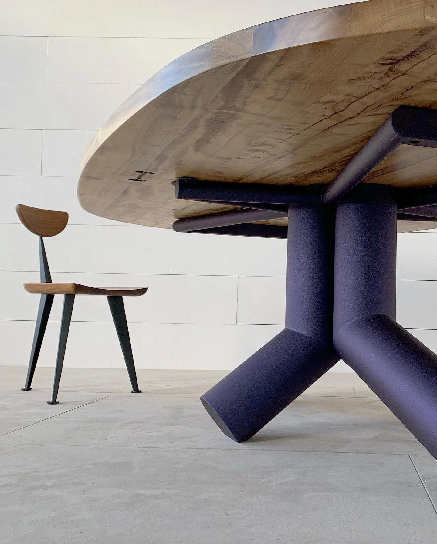 Close-up of a wooden table leg with a curved top edge and black metal supports, with a small wooden chair with black legs and a curved backrest in the background on a light-colored tiled floor with a white tiled wall.