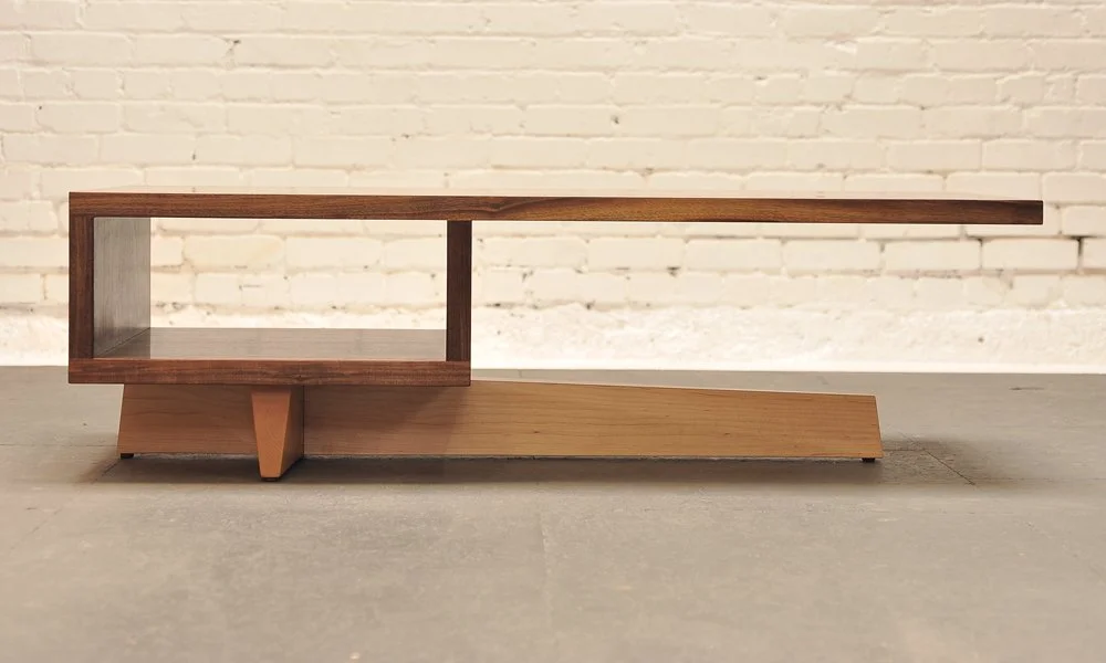 Modern wooden table with an open rectangular compartment and a sloped surface against a white brick wall.