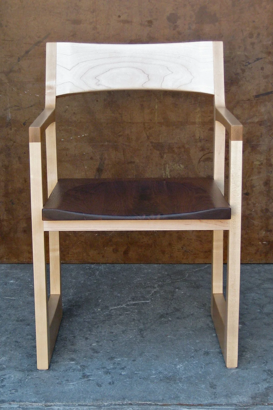 Wooden chair with a curved backrest and dark cushioned seat, set against a brown wooden wall on a concrete floor.