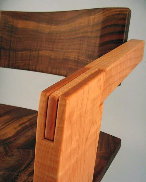Close-up of a wooden chair showing the backrest made of dark wood and the armrest made of lighter wood.