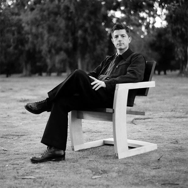 A young man sits on a modern wooden chair outdoors in a park, dressed in dark clothing, with trees in the background.