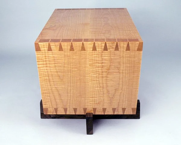 A wooden box with dovetail joints on the corners, sitting on a dark wooden stand.