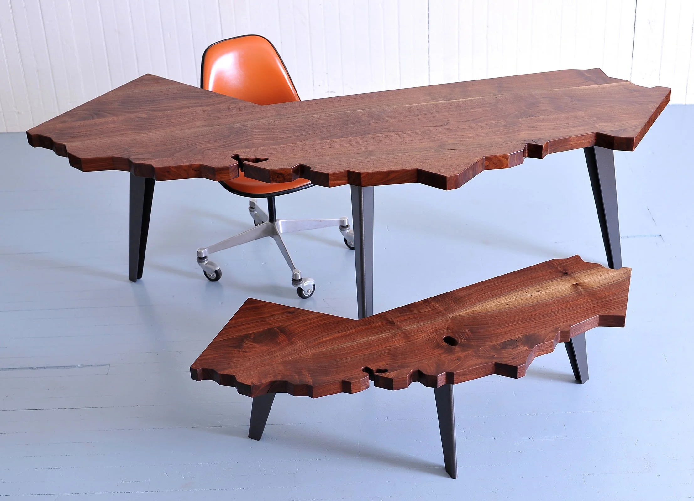 A modern wooden table with an irregular edge design, supported by black angled legs, with an orange desk chair behind it, set against a light-colored wall.