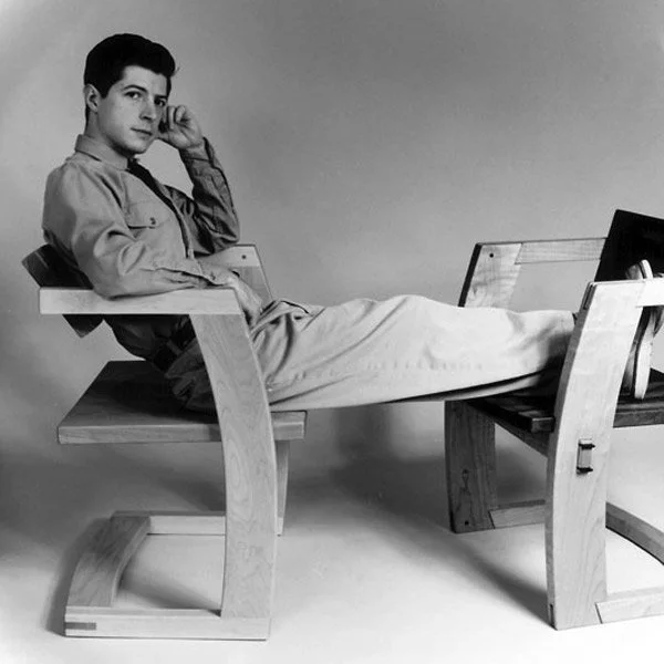 Black and white photo of a man relaxing on a wooden bench with a second bench nearby, in a studio setting.