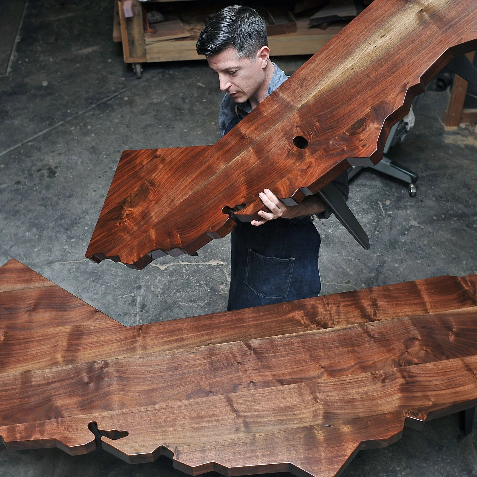 A man inspecting a large, polished wooden table with a natural edge in a woodworking shop.
