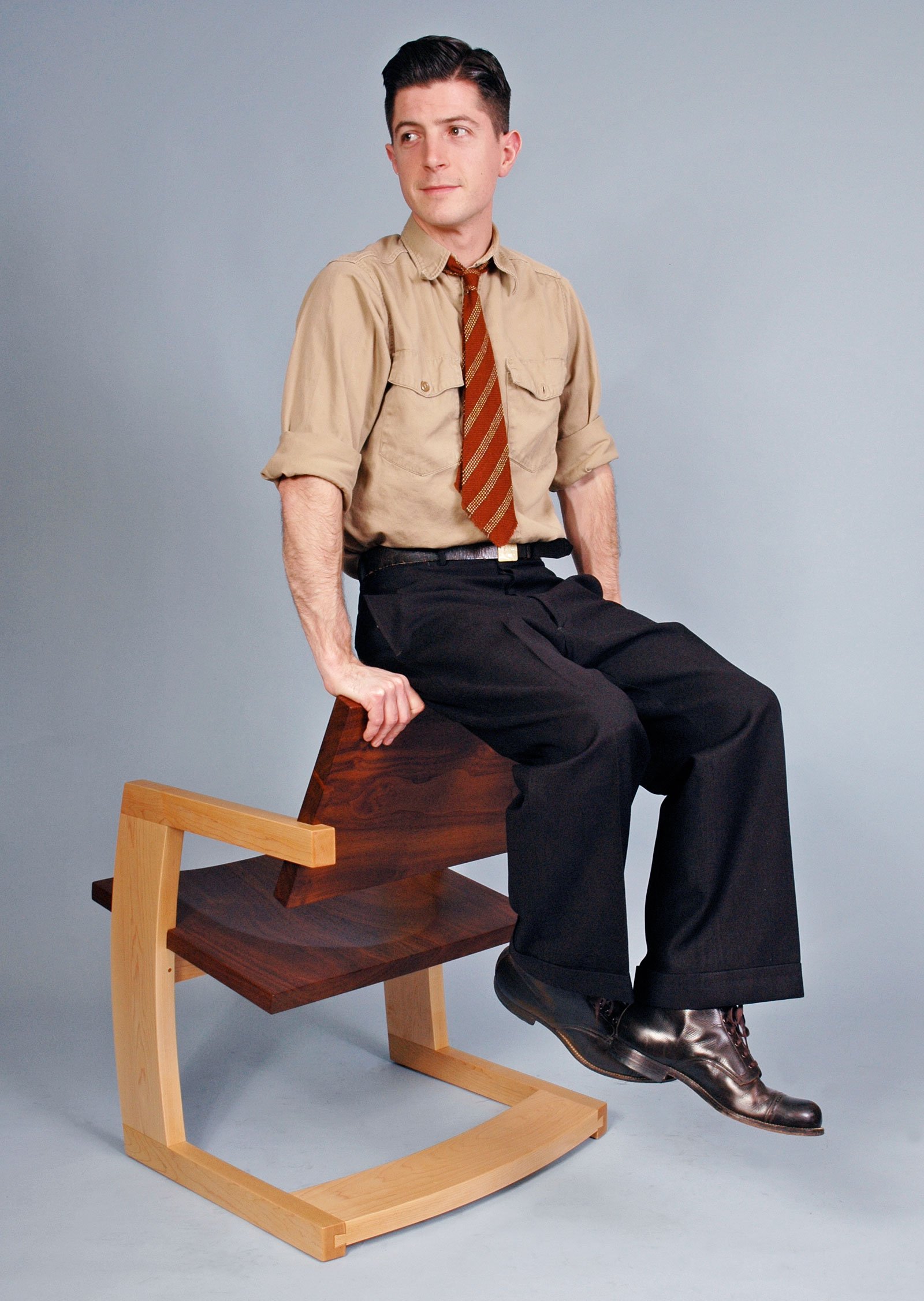 A young man with dark hair wearing a beige shirt, striped brown tie, black pants, and dress shoes sitting on a wooden chair with one leg crossed over the other, against a plain gray background.