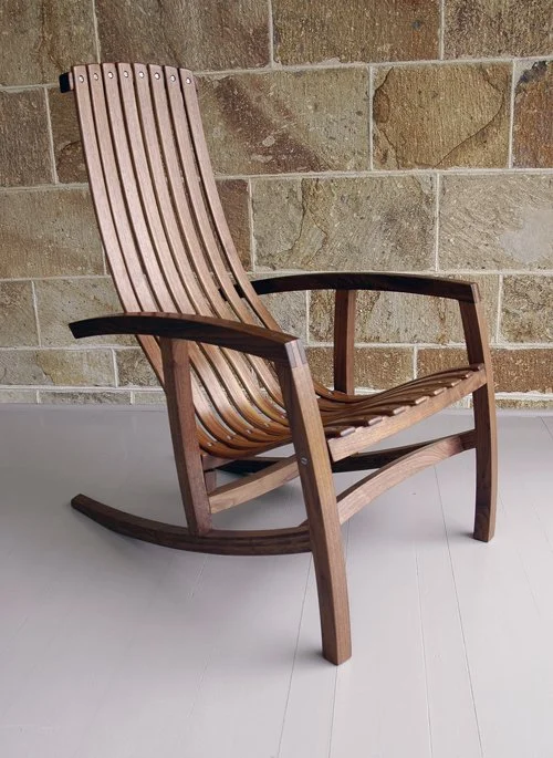 Wooden rocking chair with a slatted back and seat, positioned on a white floor against a brick wall background.